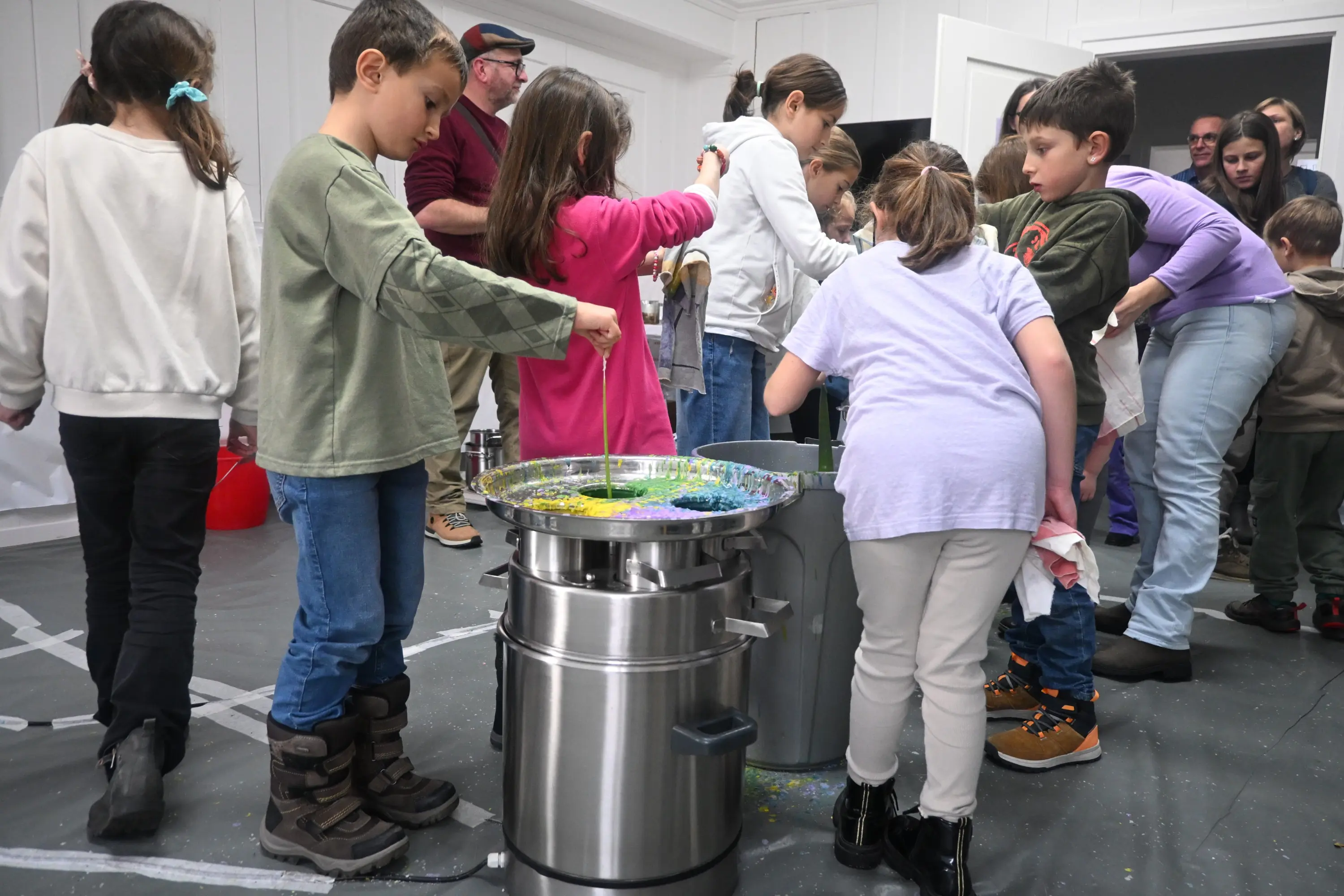 Un gruppo di bambini e adulti partecipa a un'attività creativa, mescolando colori in grandi contenitori. I bambini indossano abiti casual e sembrano divertirsi mentre