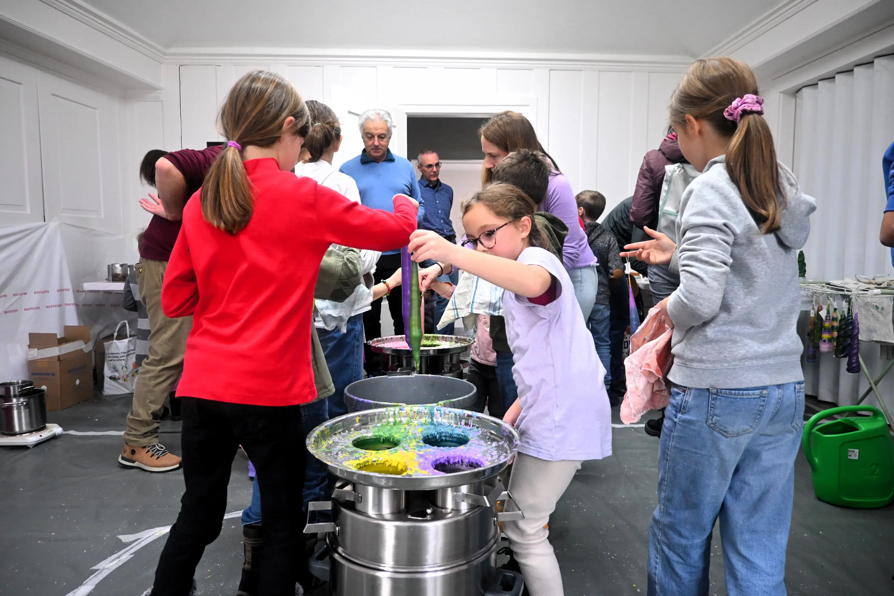 Bambini mescolano colori vivaci in grandi contenitori, mentre adulti osservano e assistono. L'ambiente è luminoso e creativo, con materiale artistico sparso attorno