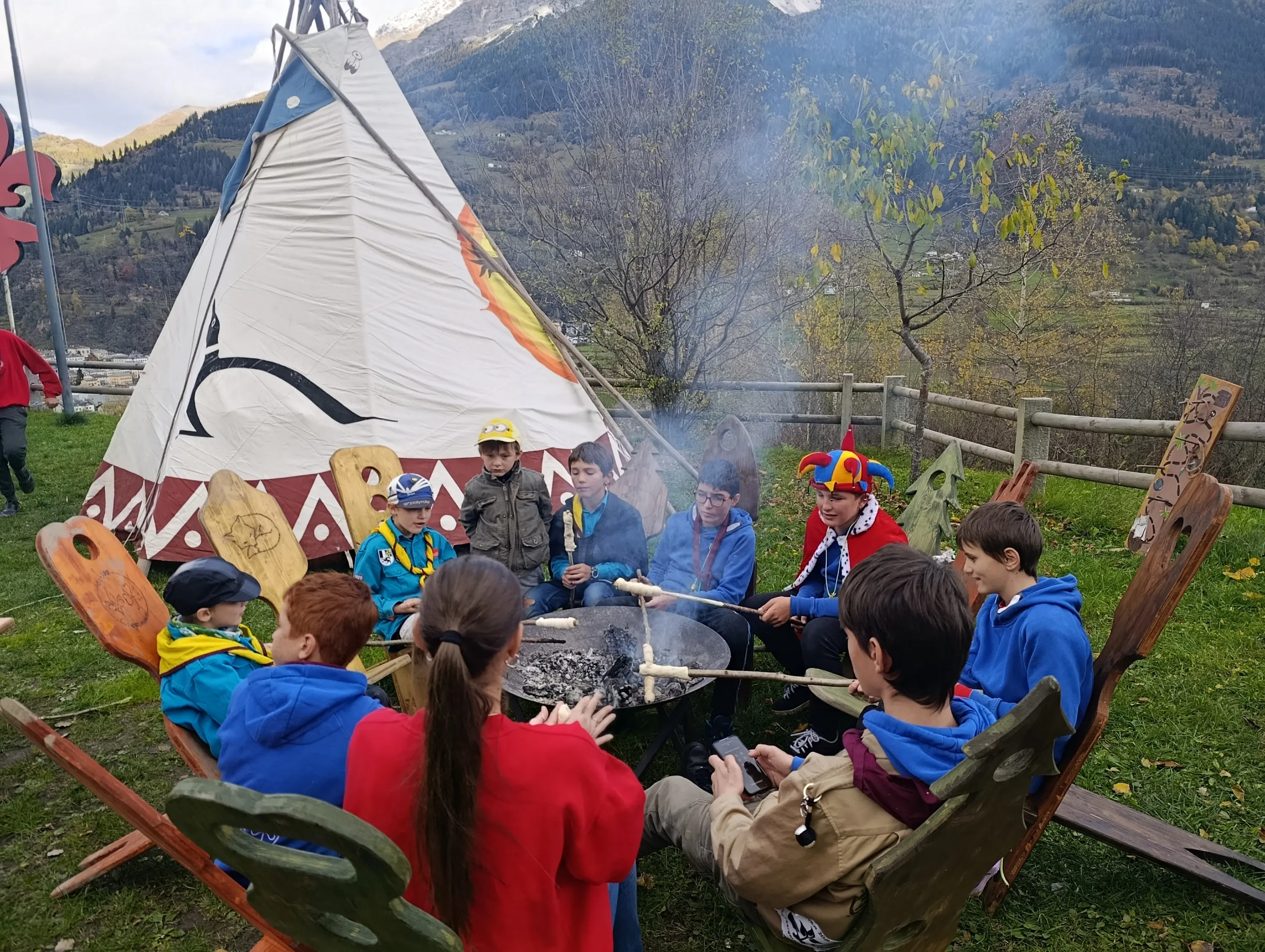Un gruppo di ragazzi seduti intorno a un falò, con un grande tendone a forma di teepee sullo sfondo. Alcuni ragazzi indossano cappelli colorati e giacche blu, mentre altri si divertono a cucinare sopra il fuoco. La natura circostante è visibile, con colline e alberi in autunno.