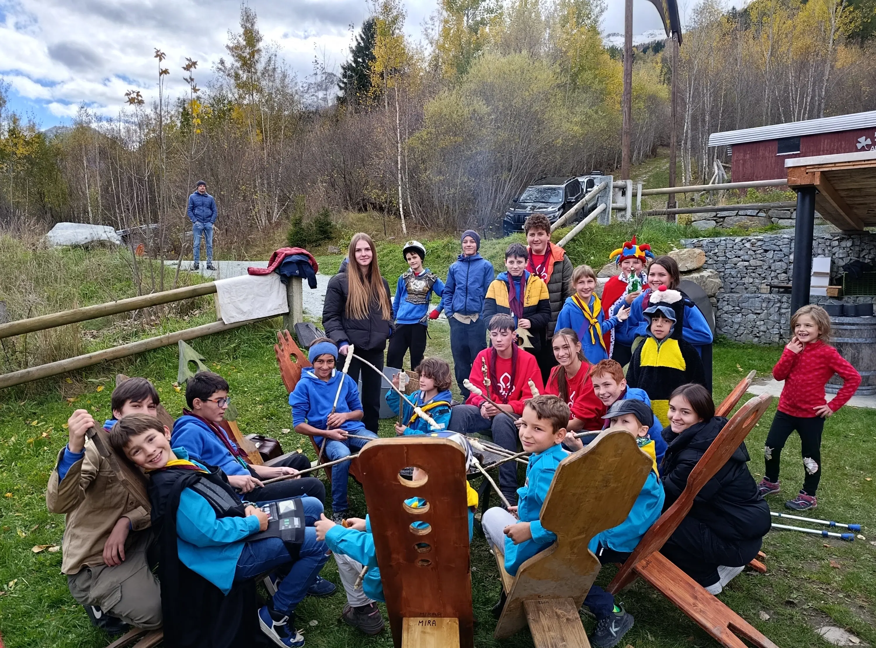 Gruppo di ragazzi e ragazze sorridenti in un campo all'aperto, alcuni vestiti in uniformi colorate, riuniti intorno a una grande struttura di legno. Sullo sfondo si vedono alberi e una struttura in legno.