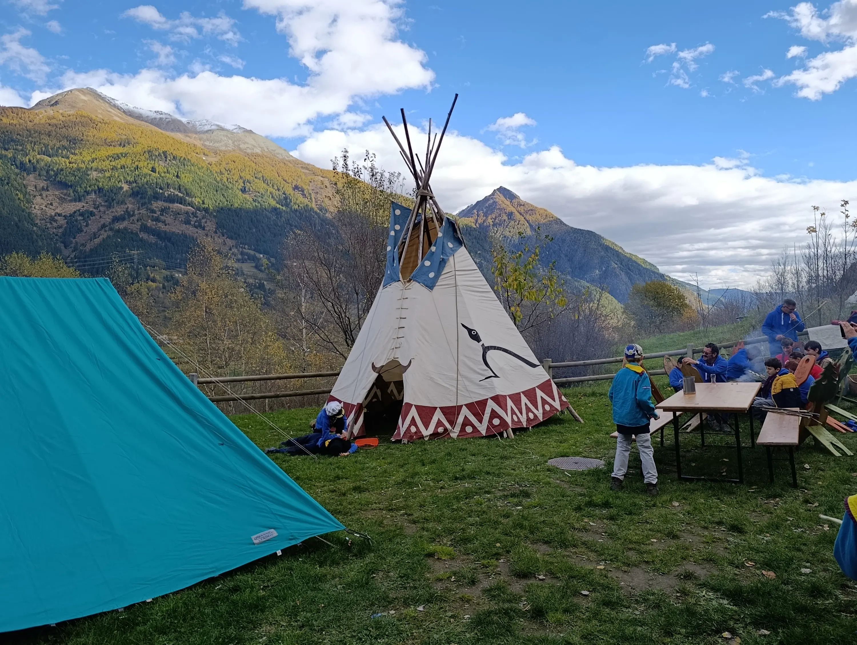 Un accampamento all'aperto con un tipì e una tenda blu, circondato da montagne. Un gruppo di persone è seduto attorno a un tavolo, mentre altri interagiscono vicino al fuoco. Il cielo è parzialmente nuvoloso e la vegetazione autunnale è visibile.