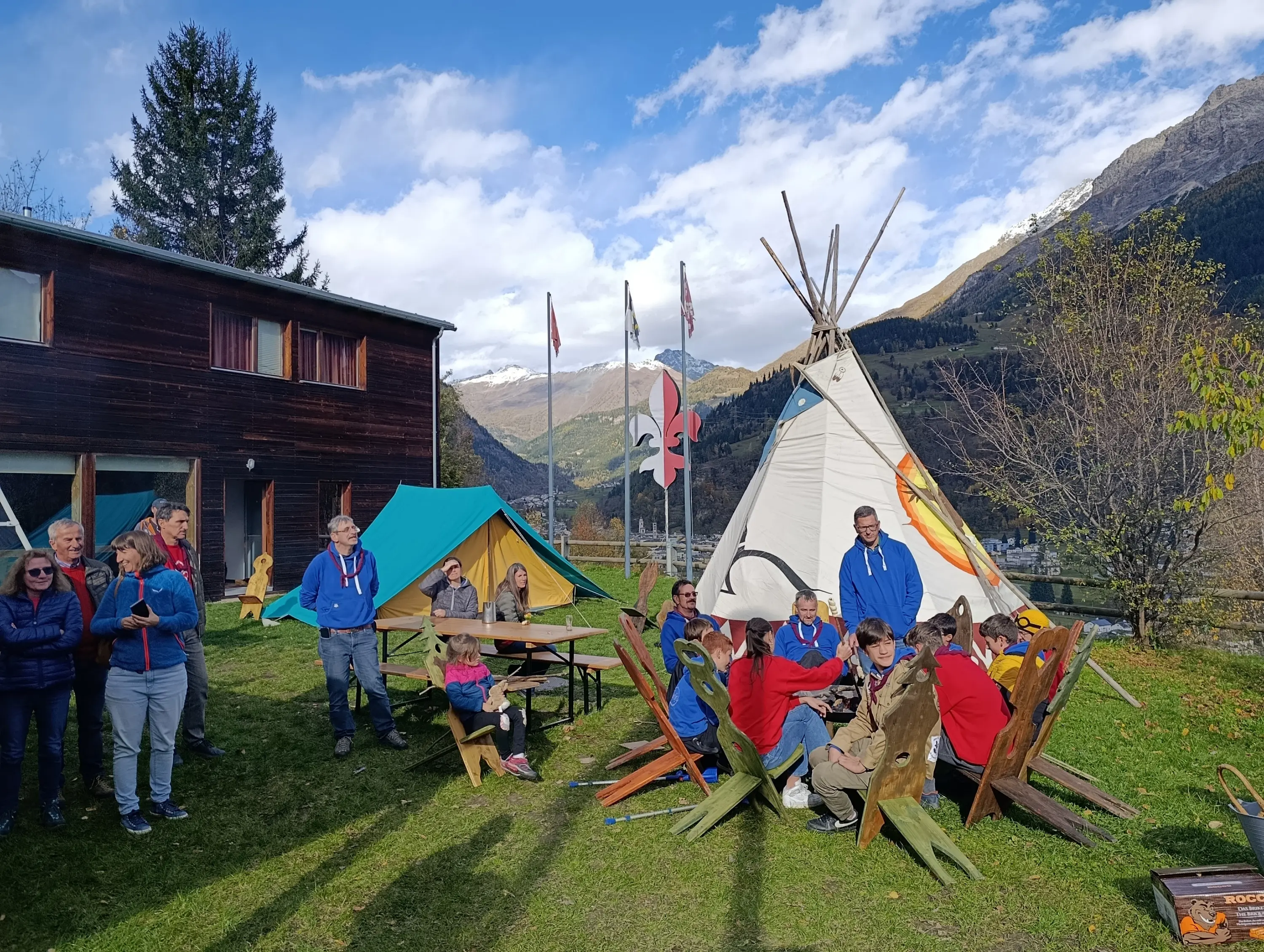Gruppo di persone riunite intorno a un fuoco all'aperto, con un grande teepee bianco sullo sfondo. Alcuni si trovano davanti a un tavolo, mentre altri stanno in piedi, tutti in un ambiente montano con bandiere e tende colorate.