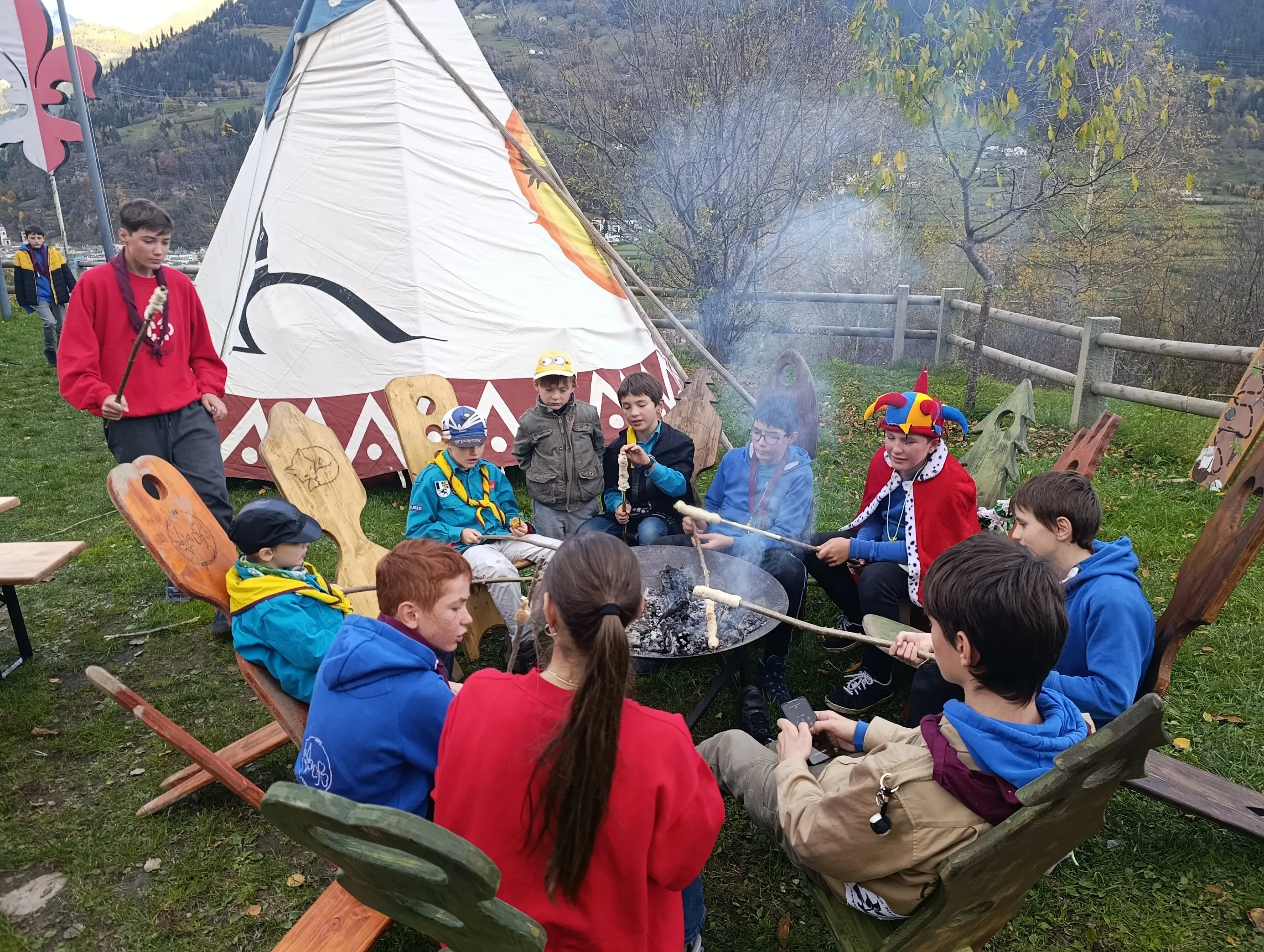 Gruppo di giovani seduti attorno a un fuoco, con un teepee sullo sfondo. Alcuni indossano cappelli colorati e mostrano sorrisi. L'ambientazione è naturale, con alberi e montagne in lontananza.