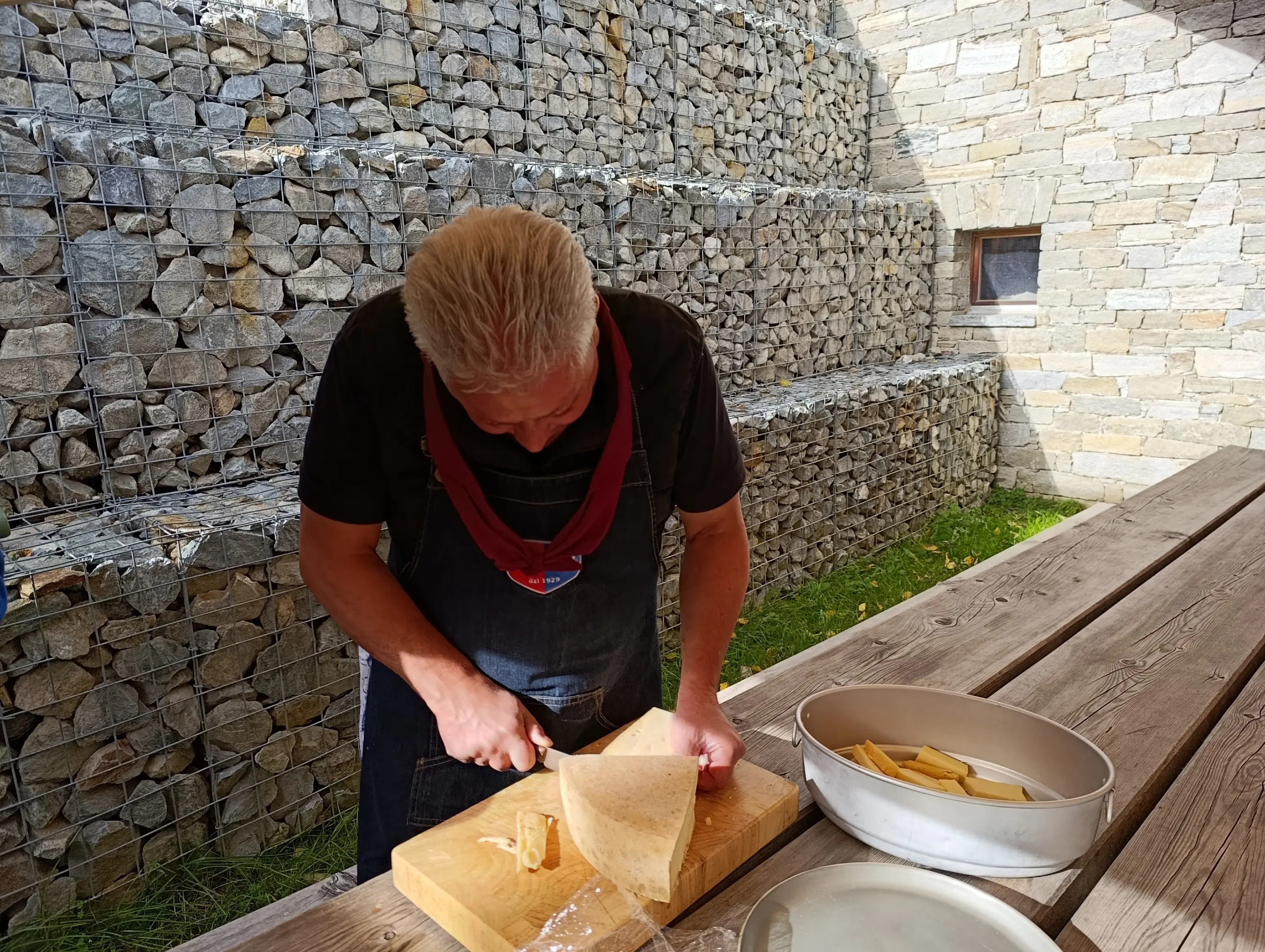 Un uomo sta affettando un grande pezzo di formaggio su un tagliere, all'aperto, con una parete di pietre sullo sfondo. Indossa un grembiule e si concentra sul lavoro.