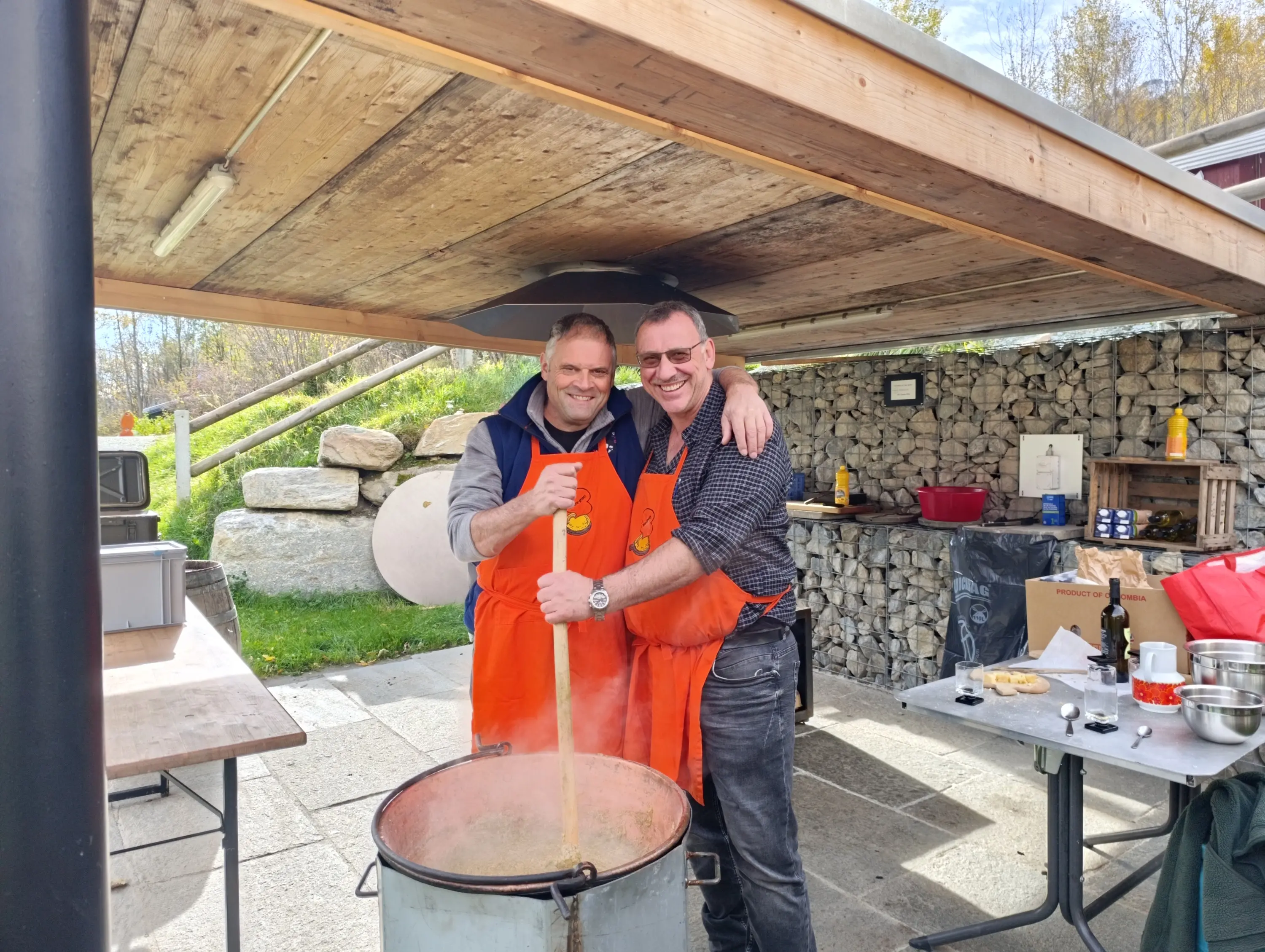 Due uomini sorridenti indossano grembiuli arancioni mentre mescolano un grande pentolone all'aperto. Sullo sfondo, ci sono rocce e attrezzature da cucina. È una giornata di sole.