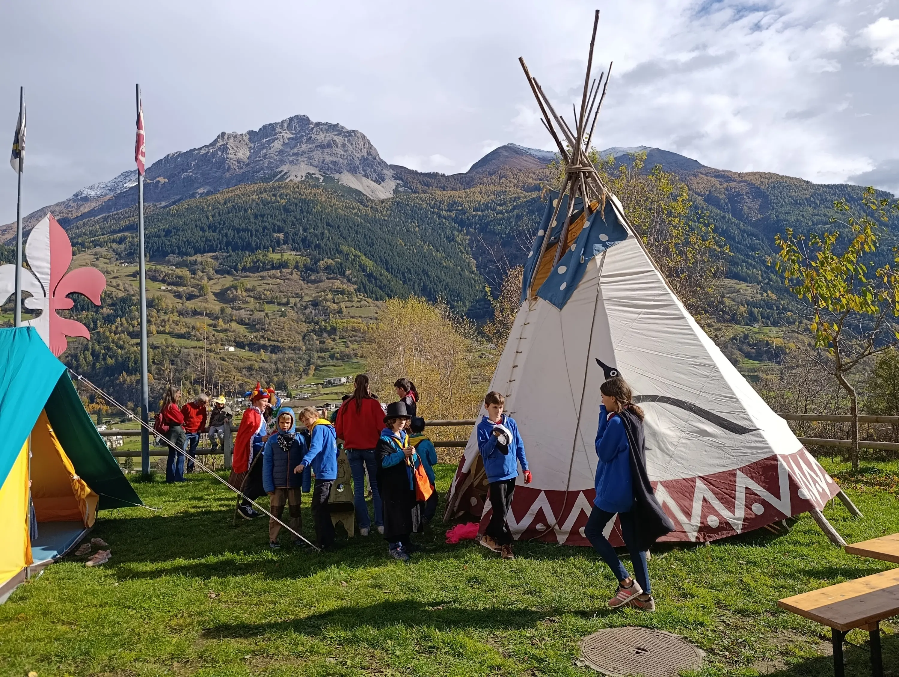 Gruppo di bambini e adulti vicino a un grande teepee, circondato da montagne e un clima autunnale sereno. Alcune bandiere sventolano in lontananza.