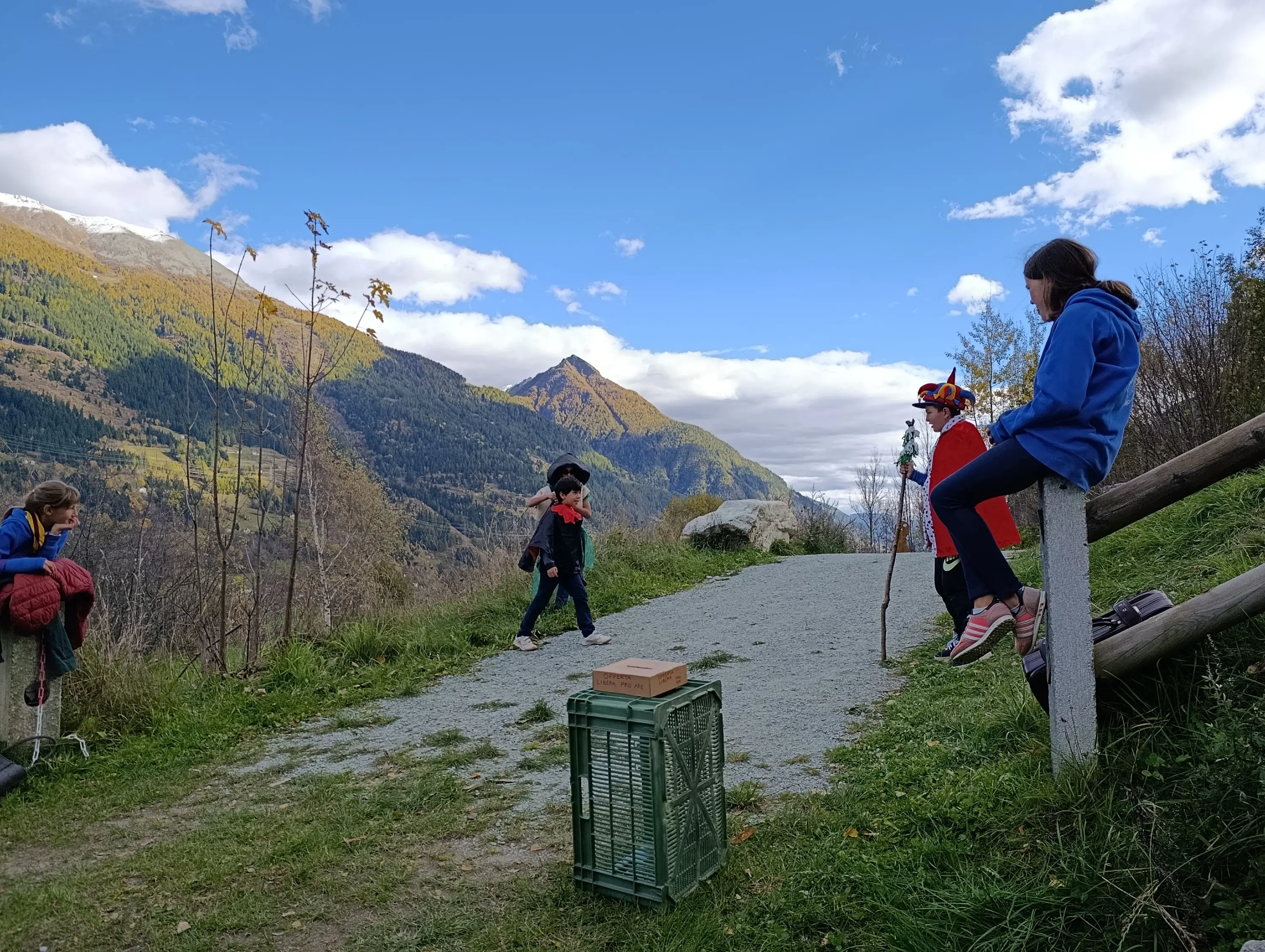 Gruppo di bambini in un paesaggio montano, alcuni seduti su un muretto e altri in piedi lungo un sentiero. Si vedono alberi e montagne sullo sfondo, con un cielo blu e nuvole. Una cassa verde è posizionata a terra.