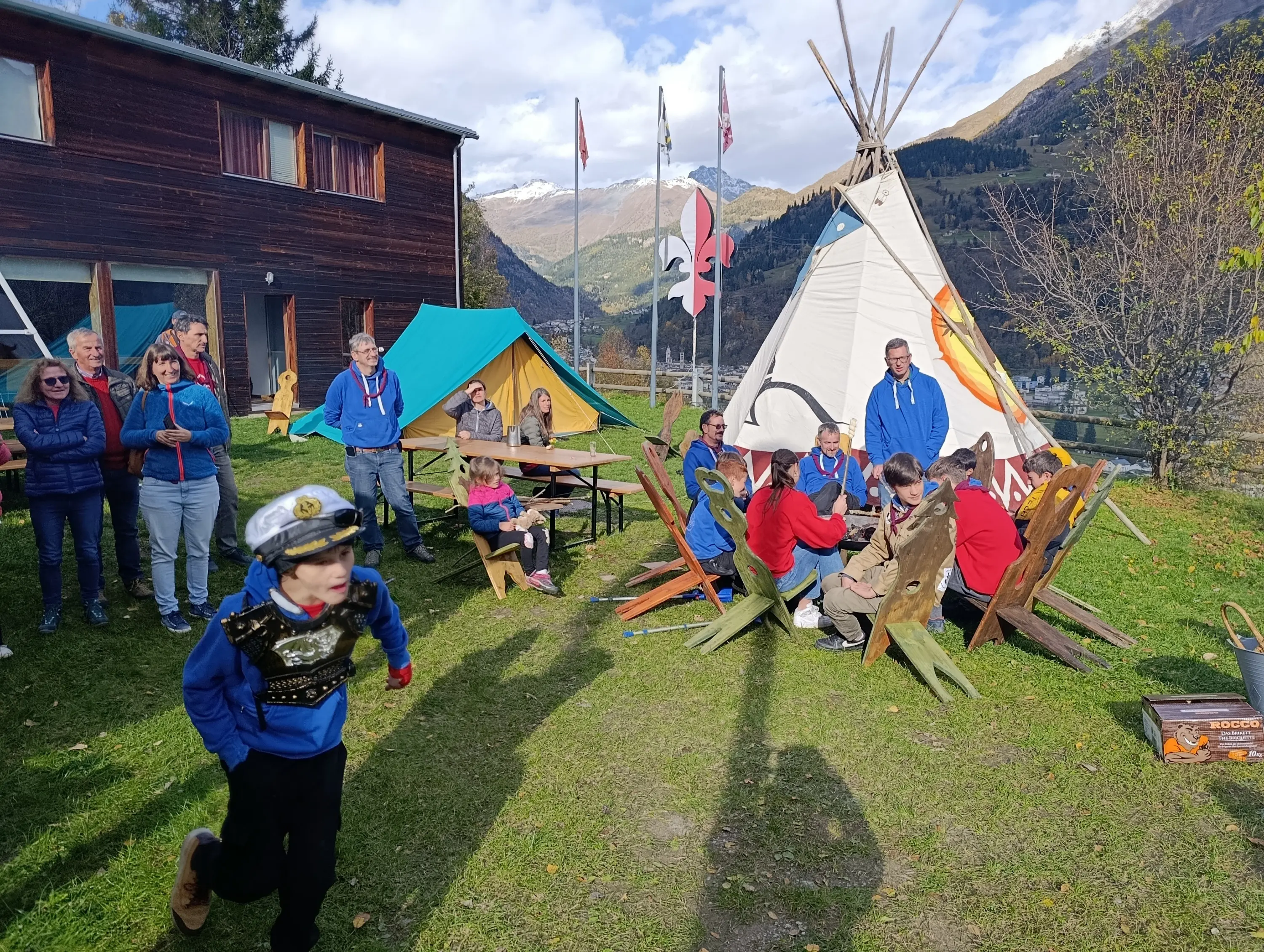 Un gruppo di persone si riunisce all'aperto, vicino a tende e un grande tendone indiano, circondato da montagne. Alcuni adulti osservano mentre dei bambini giocano, uno dei quali indossa un costume da capitano.