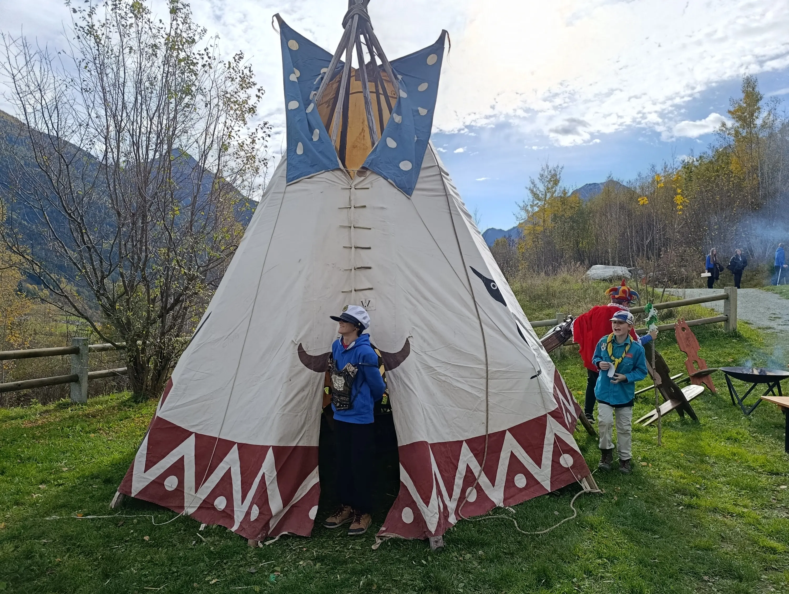 Due bambini si trovano accanto a un grande teepee decorato, immersi in un paesaggio autunnale. Uno dei bambini indossa un cappello e sembra impegnato in un'attività, mentre un altro osserva. Gli alberi intorno mostrano foglie gialle e l'aria è serena.