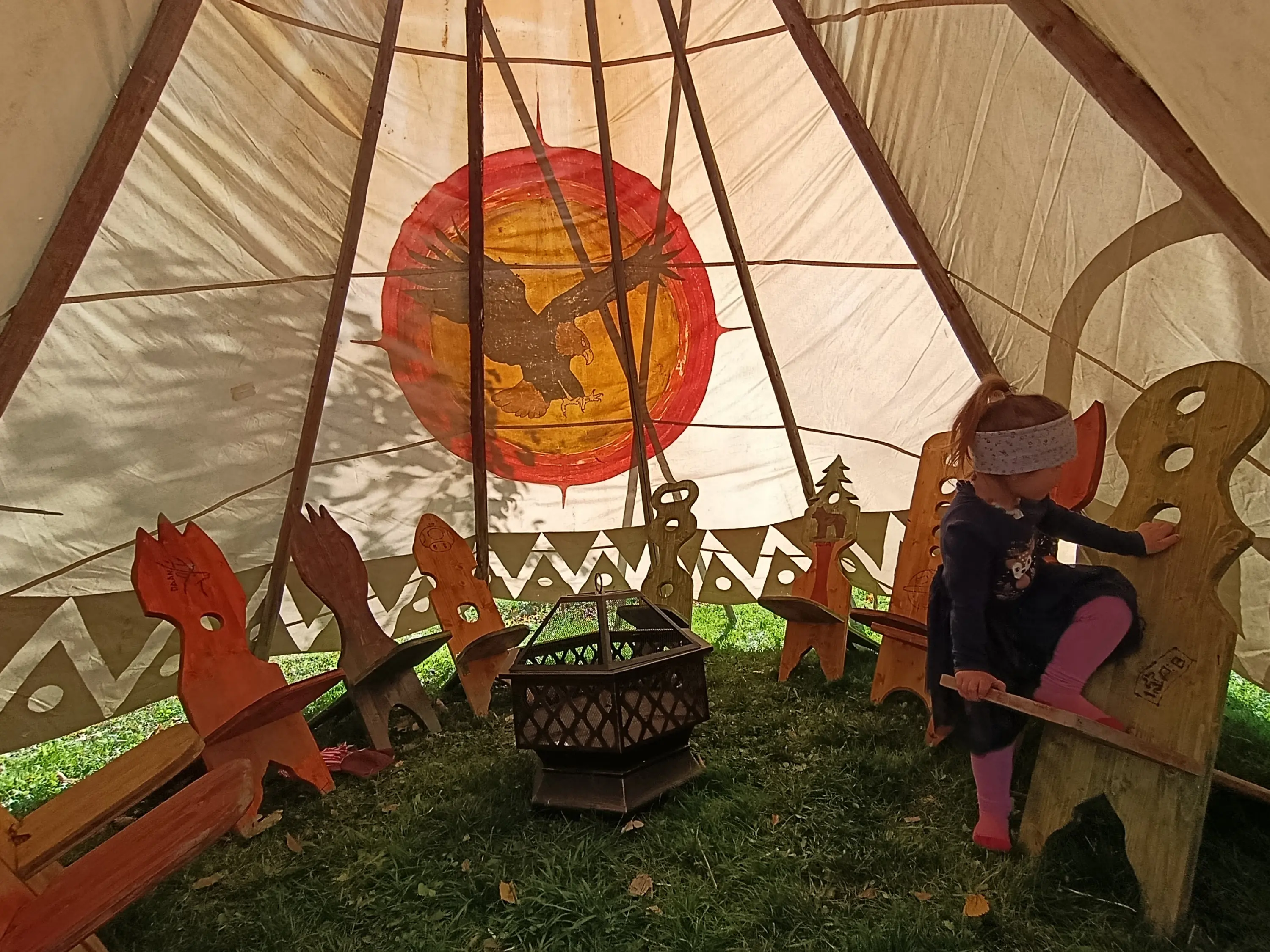 Interno di un tendone con panchine di legno decorate e un bambino che gioca. Al centro c'è un camino e sul retro si vede un disegno di un animale. La luce filtra attraverso la tela.