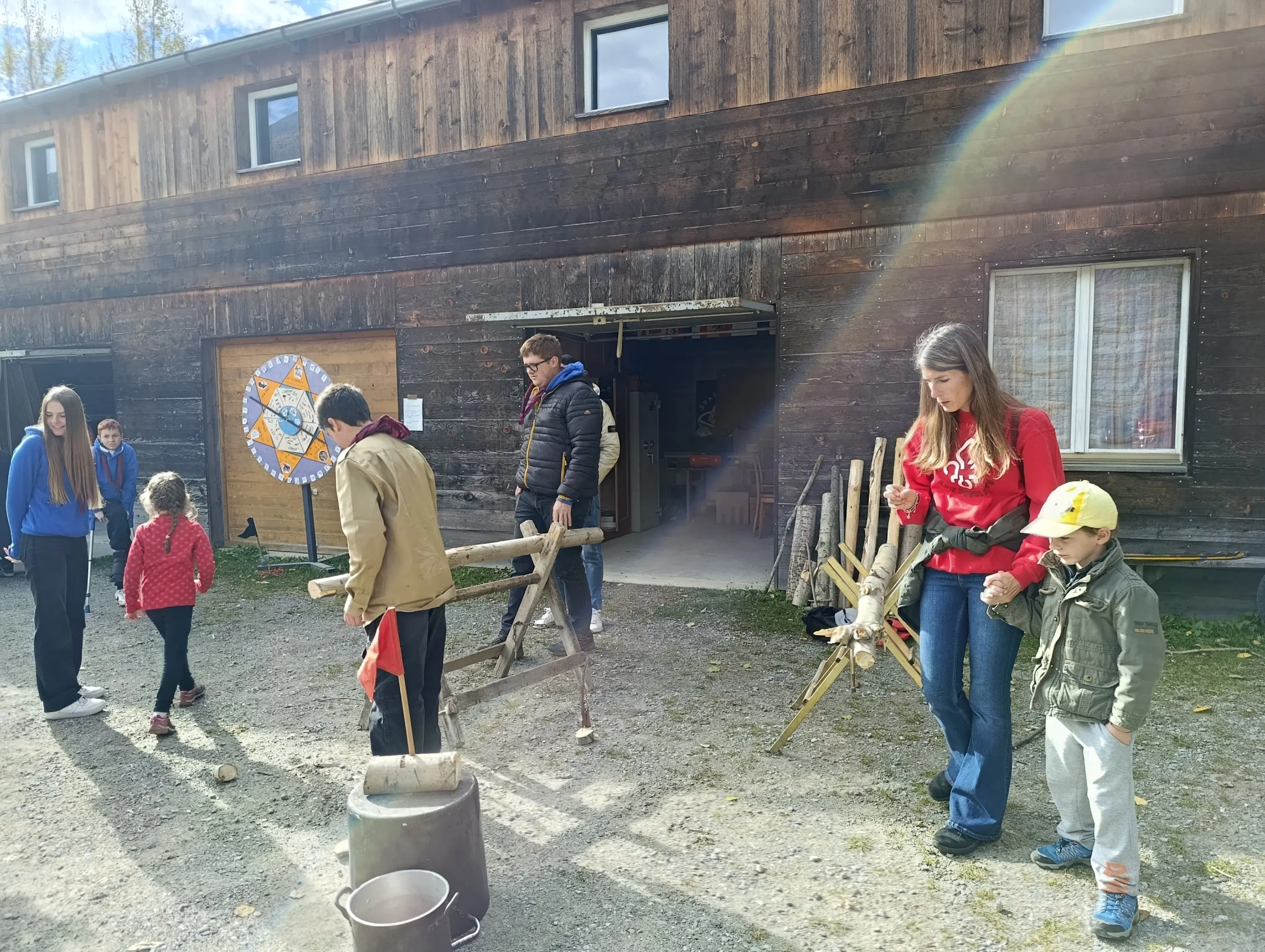 Attività all'aperto in un ambiente rurale. Un gruppo di persone, tra cui adulti e bambini, utilizza strumenti di legno mentre un edificio in legno si trova sullo sfondo. Alcuni sorridono e interagiscono tra loro, mentre la luce del sole illumina la scena.