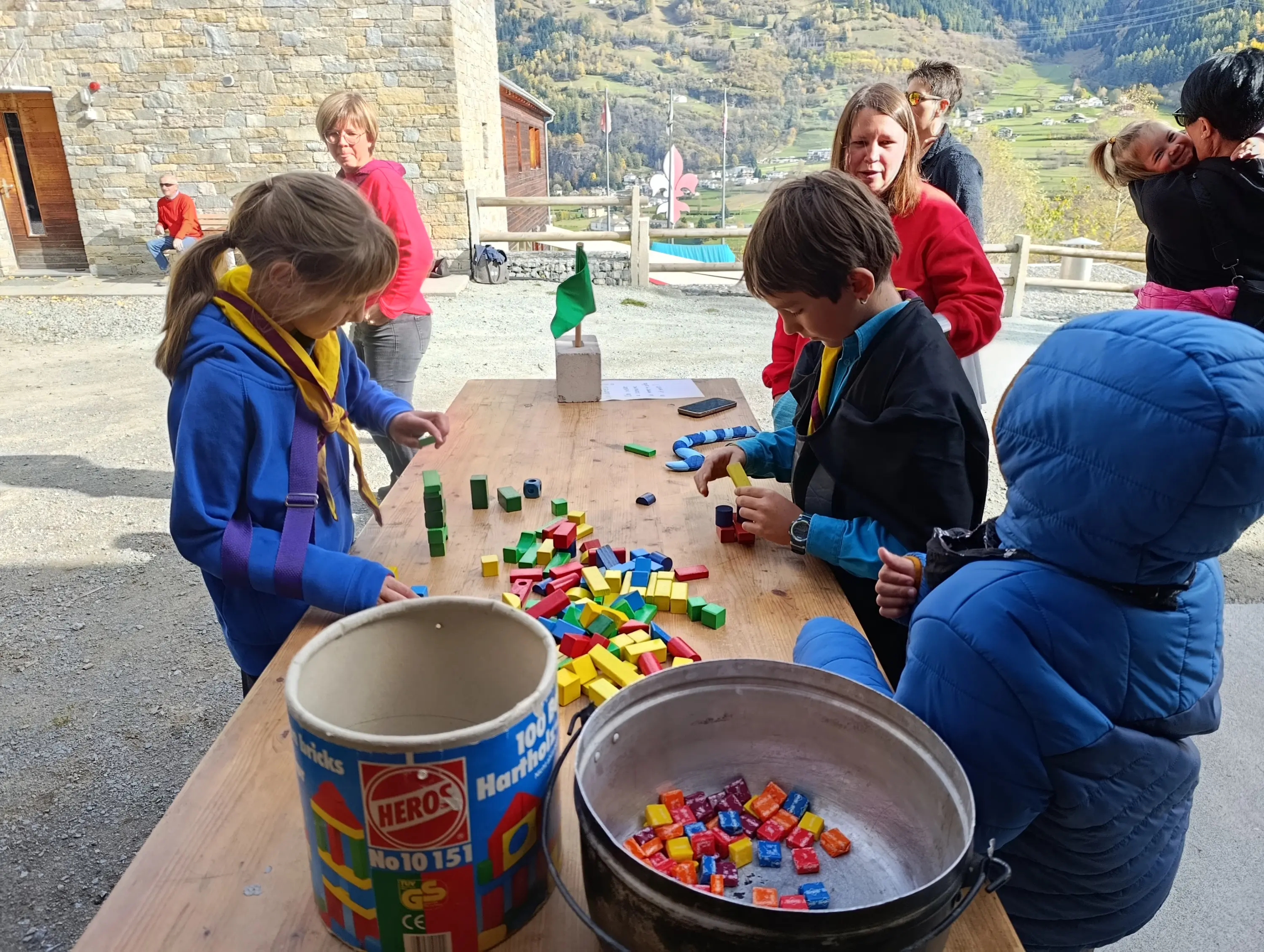 Bambini che giocano con blocchi colorati su un tavolo all'aperto, con montagne sullo sfondo. Alcuni adulti interagiscono con i ragazzi, creando un'atmosfera vivace e divertente.