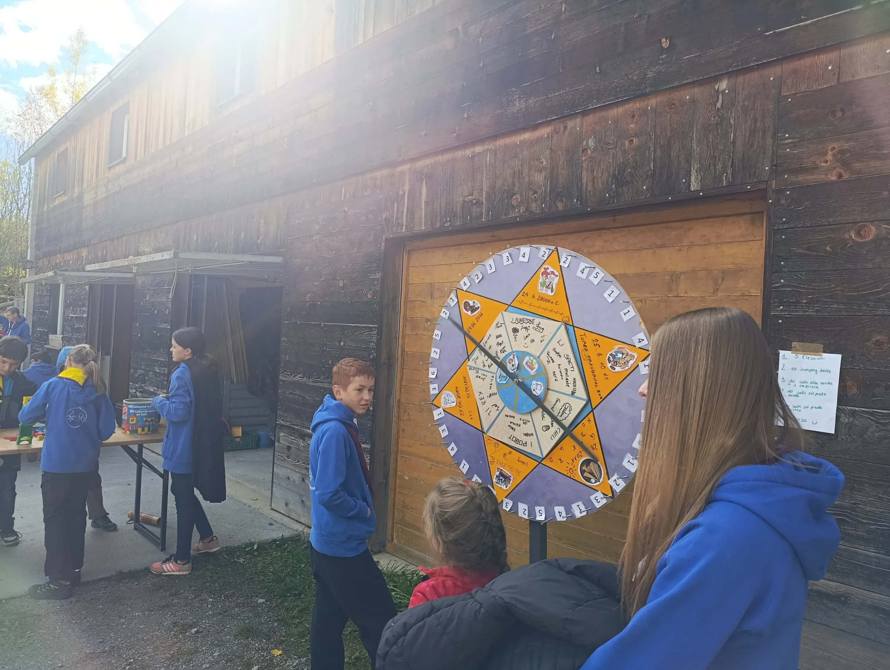 Un gruppo di bambini e ragazzi interagiscono con una grande ruota della fortuna all'aperto, davanti a un edificio di legno. Alcuni bambini sono impegnati in giochi mentre altri osservano. All'orizzonte, il cielo è parzialmente nuvoloso.