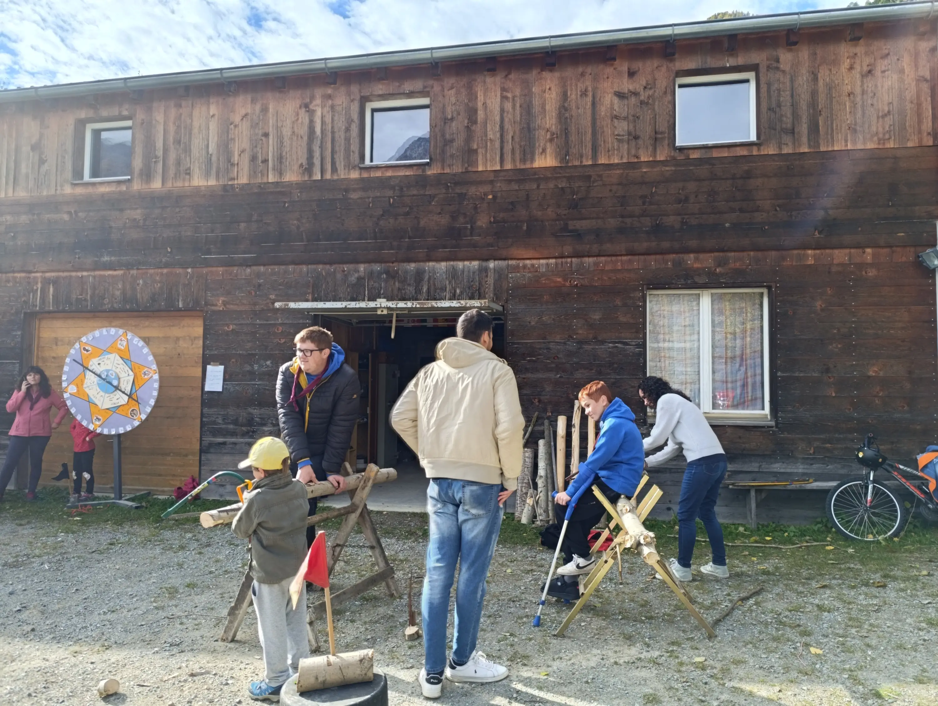 Gruppo di persone all'esterno di un edificio di legno, mentre alcuni lavorano su strutture di legno. Un bambino osserva, mentre altri adulti sono intenti nelle attività. Nel background si vede un'altra persona che scatta foto.