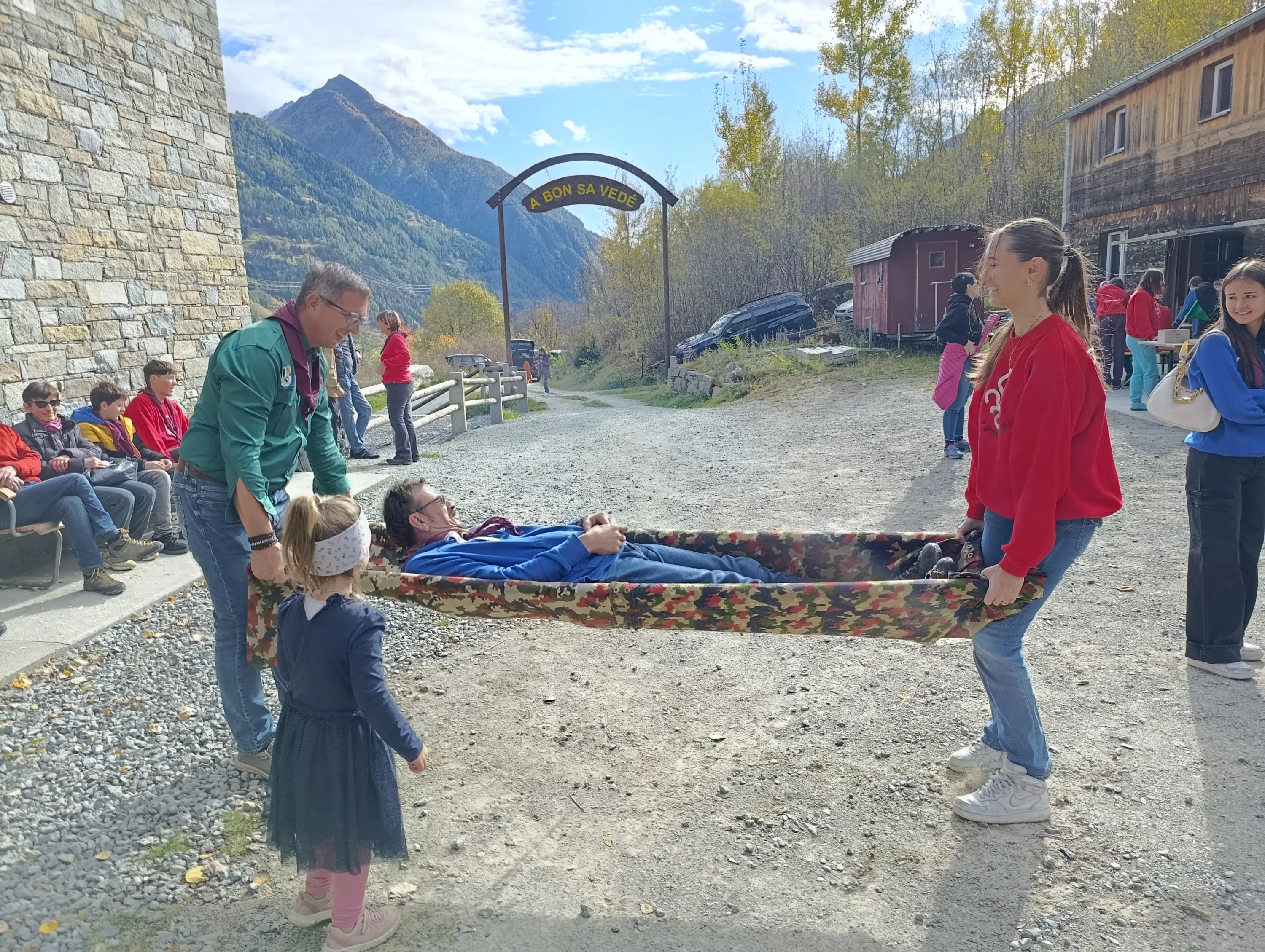 Un gruppo di persone aiuta un ragazzo disteso su una barella improvvisata, mentre una bambina guarda. Sullo sfondo si intravedono le montagne e altre persone sedute.