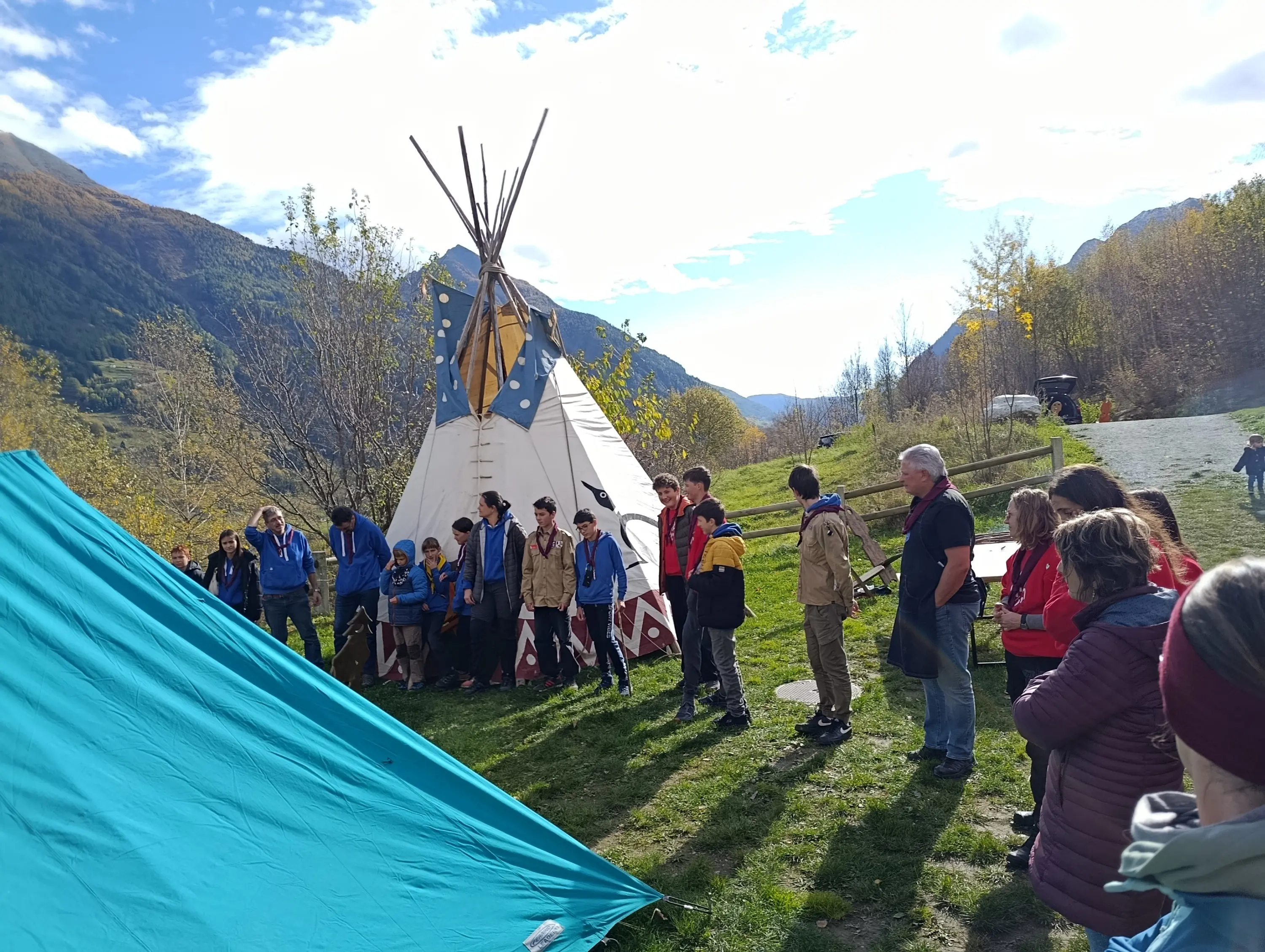 Un gruppo di persone si trova all'aperto vicino a un grande teepee. Alcuni partecipanti ascoltano, mentre altri sembrano conversare. Sullo sfondo, si vedono alberi autunnali e montagne, sotto un cielo parzialmente nuvoloso.