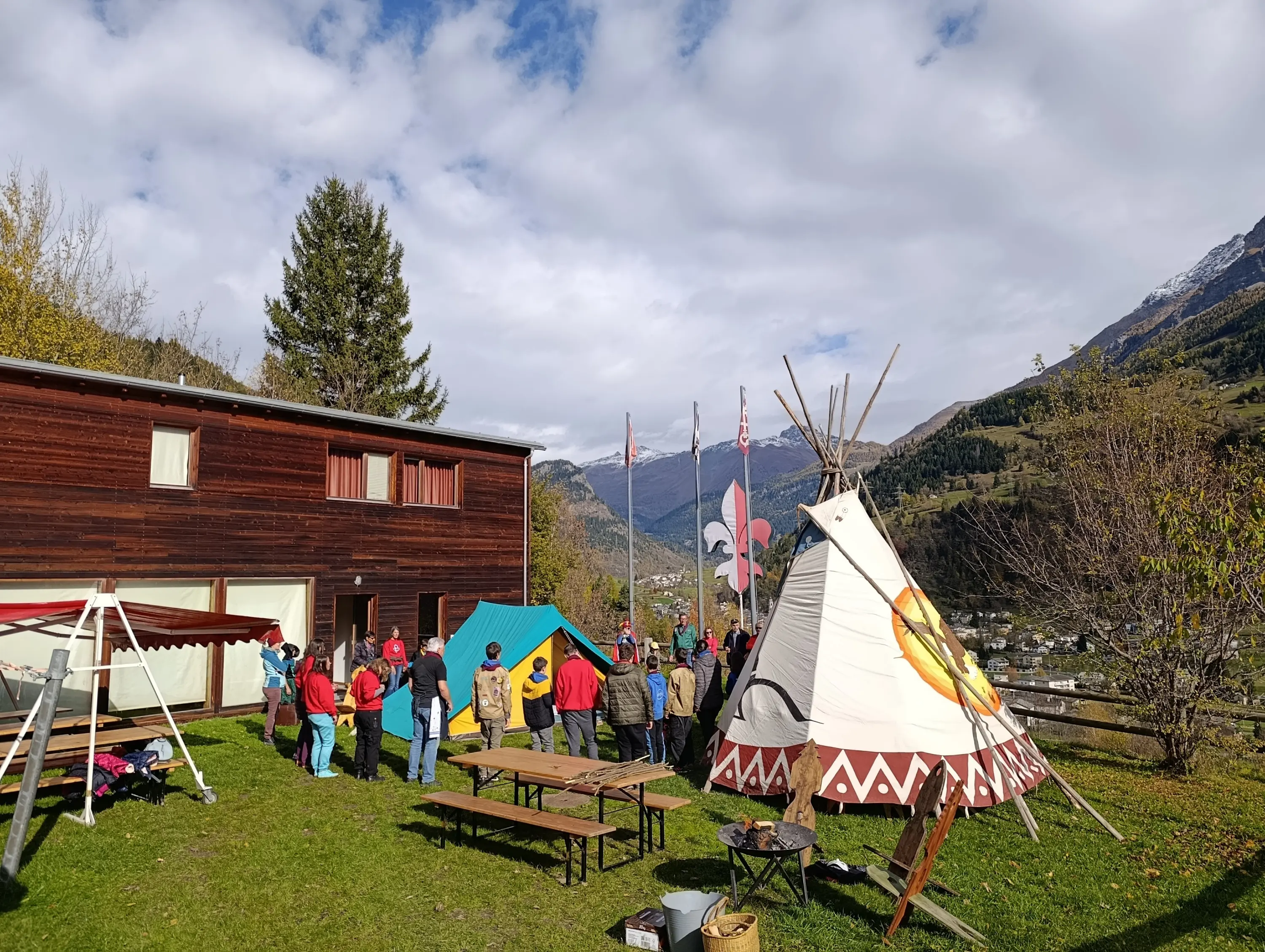 Un gruppo di persone si riunisce all'aperto vicino a tende e un teepee, con un paesaggio montano sullo sfondo. Il cielo è parzialmente nuvoloso e ci sono alberi circostanti.