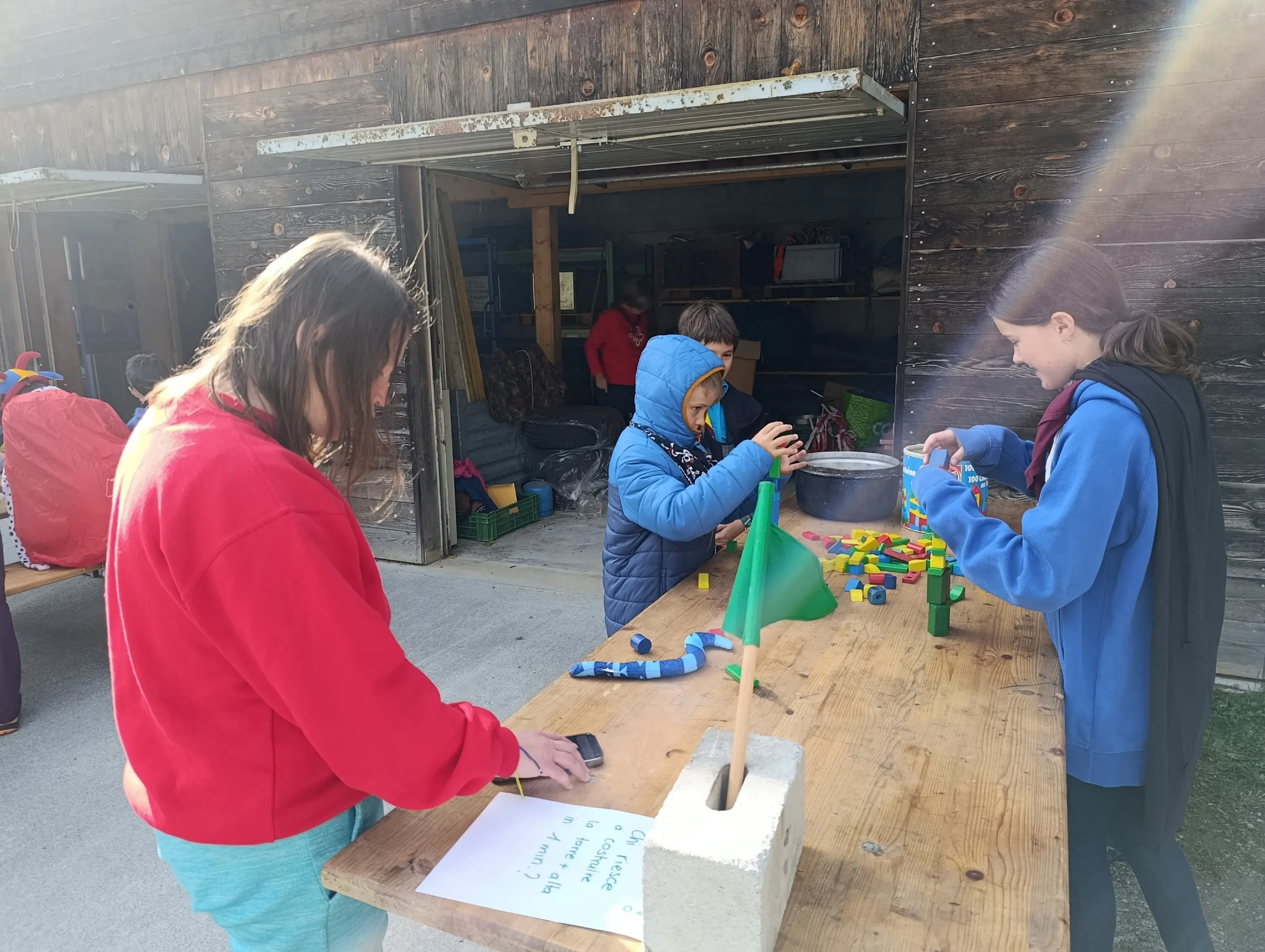 Gruppo di ragazzi che giocano all'aperto, costruendo con materiali colorati su un tavolo di legno. Sullo sfondo si vedono altre persone e una struttura in legno.