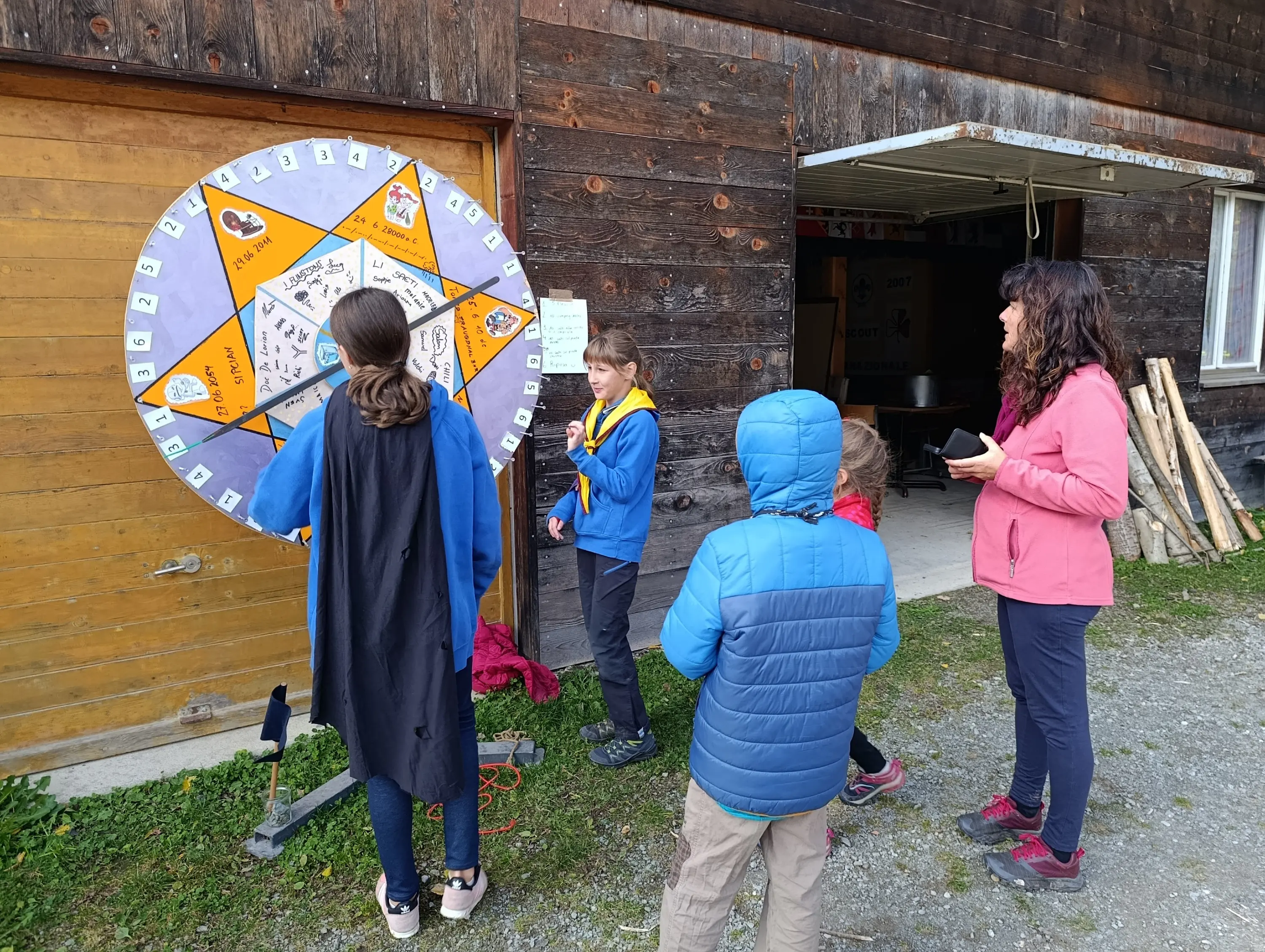 Gruppo di bambini e un adulto interagiscono con una ruota della fortuna, con diversi simboli e testi, all'interno di un ambiente rurale. Due bambini indossano giacche colorate, mentre uno ha un mantello. Si vedono anche alcuni materiali in legno sullo sfondo.