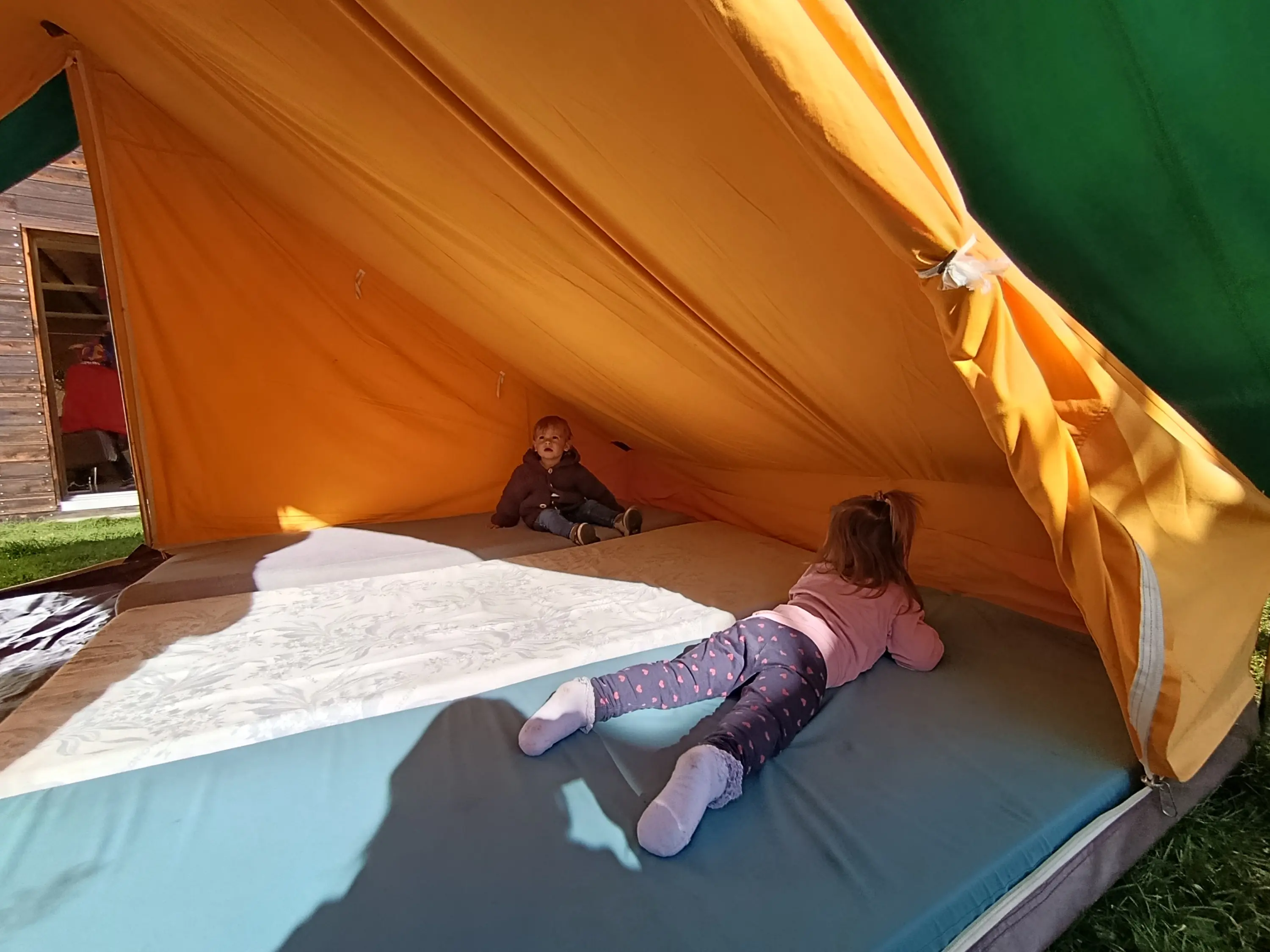 Due bambini sdraiati all'interno di una tenda colorata, immersi in un ambiente all'aperto. Un bambino è sdraiato sulla schiena, mentre l'altra bimba si muove verso di lui.