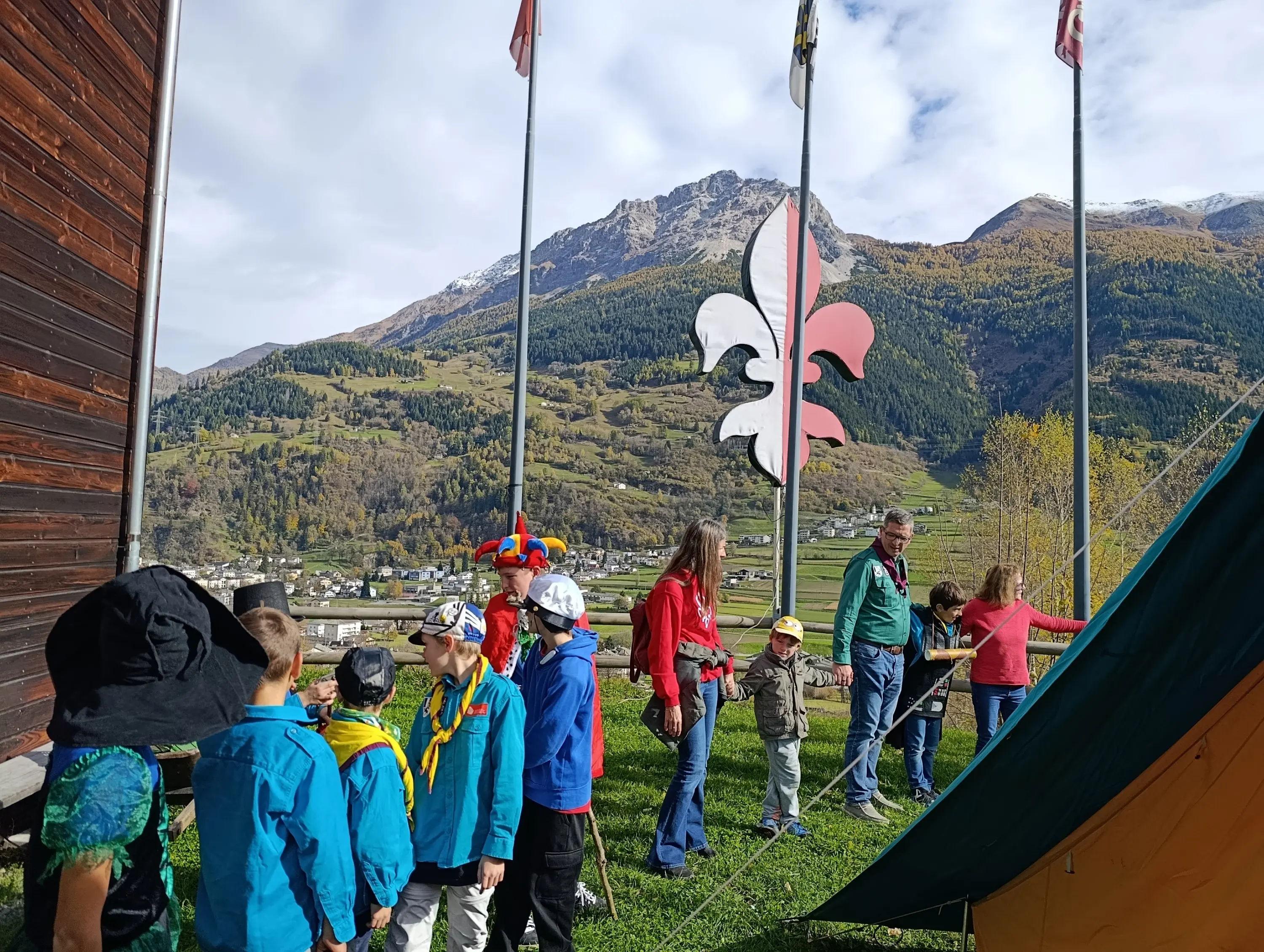 Gruppo di bambini in abbigliamento colorato mentre partecipano a un'attività all'aperto, con bandiere e montagne sullo sfondo. Una donna in rosso interagisce con i ragazzi vicino a una tenda.