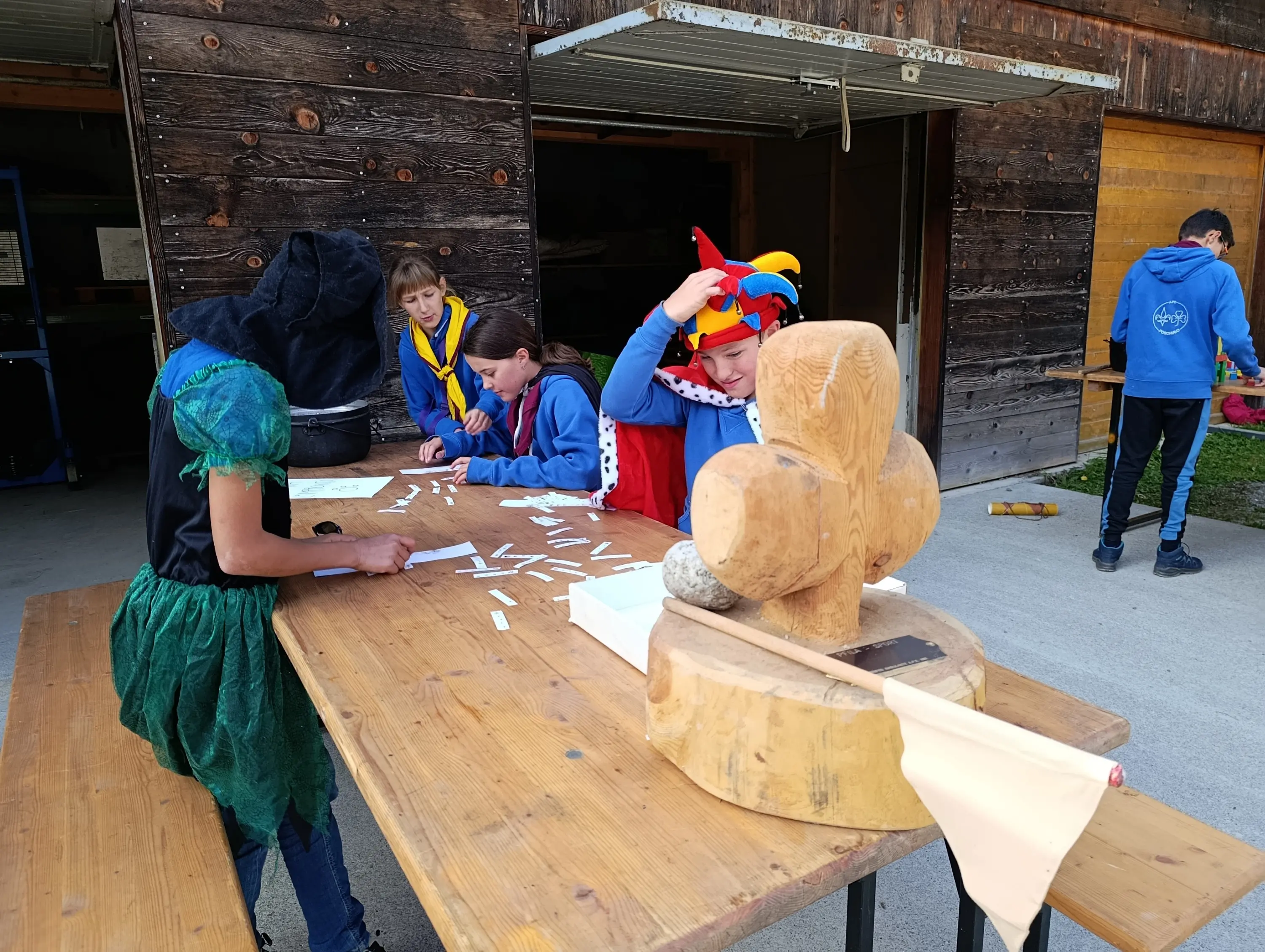 Gruppo di bambini in abiti colorati mentre partecipano a un'attività creativa all'aperto. Un tavolo di legno è decorato con diversi oggetti e materiali. Sullo sfondo, una struttura in legno.