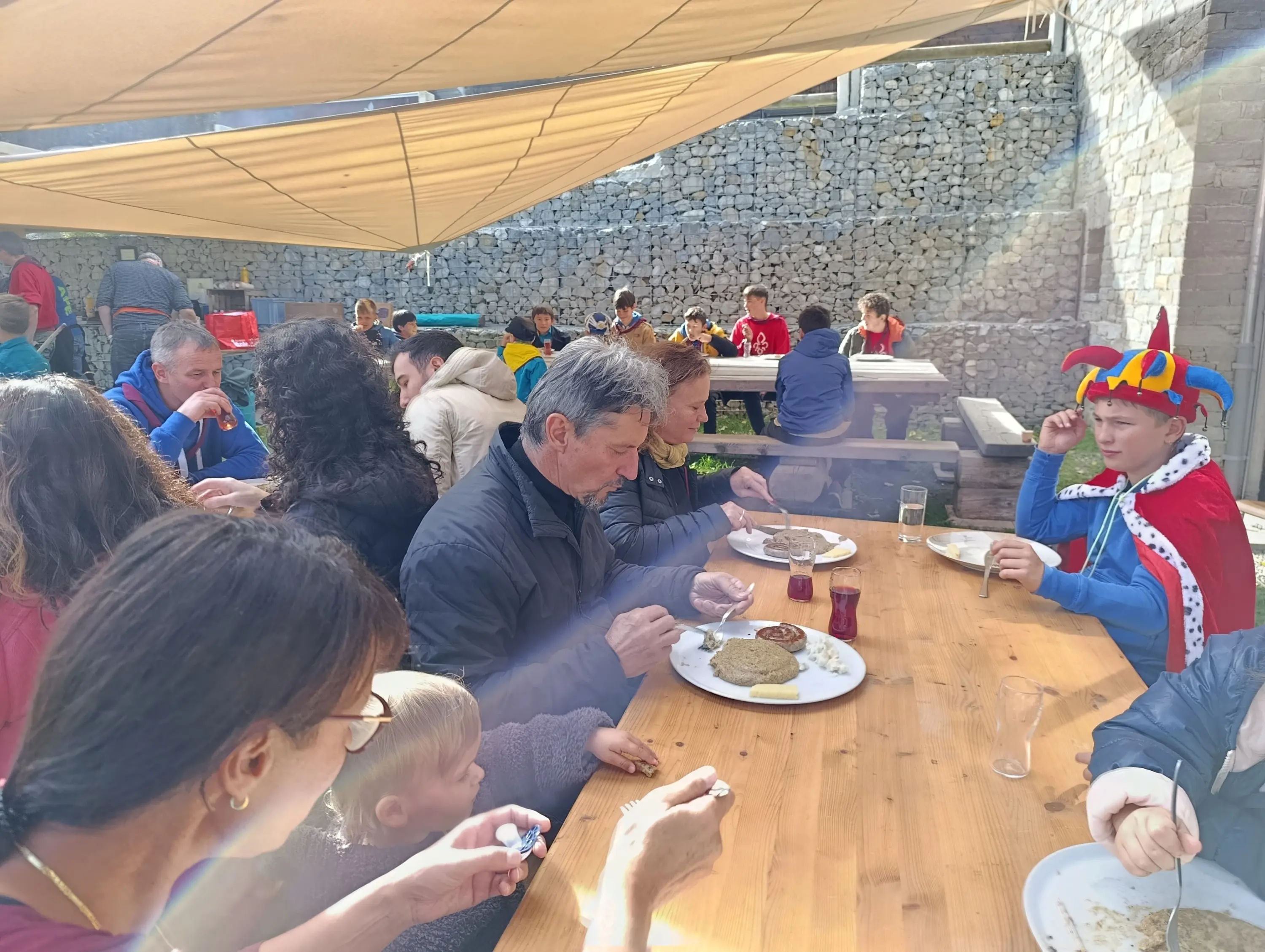 Un gruppo di persone sedute a un lungo tavolo all'aperto, mentre mangiano e chiacchierano. Alcuni bambini e adulti indossano costume, in un'atmosfera festosa. Una tenda color crema fornisce ombra sul tavolo.