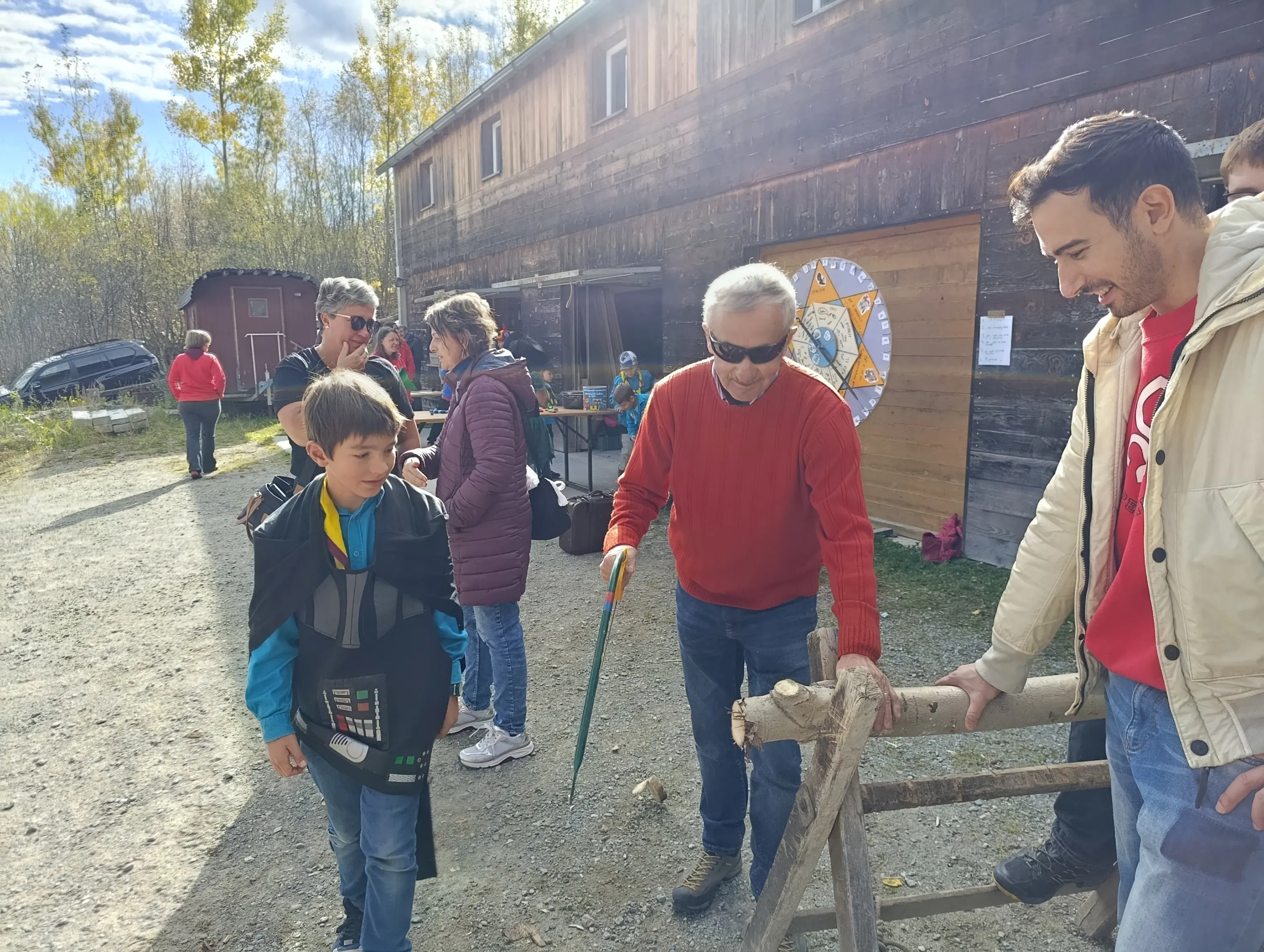 Gruppo di persone all'aperto in un'area di legno, con un uomo che tiene un bastone mentre interagisce con un ragazzo vestito in modo casuale. Altre persone osservano e ci sono diverse attività in background. Il clima è soleggiato e sereno.