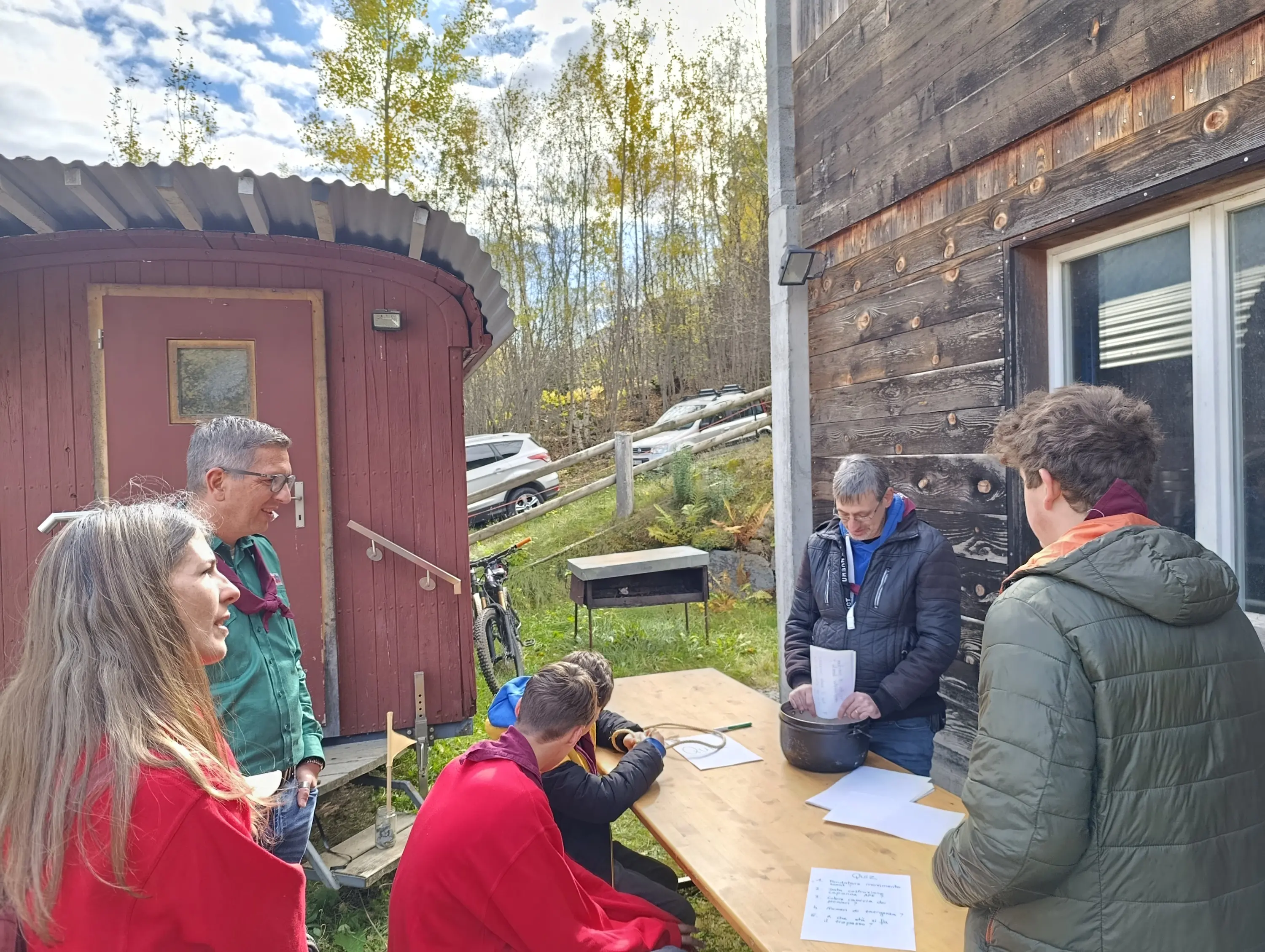 Gruppo di persone intorno a un tavolo all'aperto. Due persone sono sedute a un tavolo mentre altre tre sono in piedi: una persona sta mescolando qualcosa in una pentola, mentre gli altri osservano. Sullo sfondo si vede una piccola struttura rossa e alberi che stanno cambiando colore autunnale.