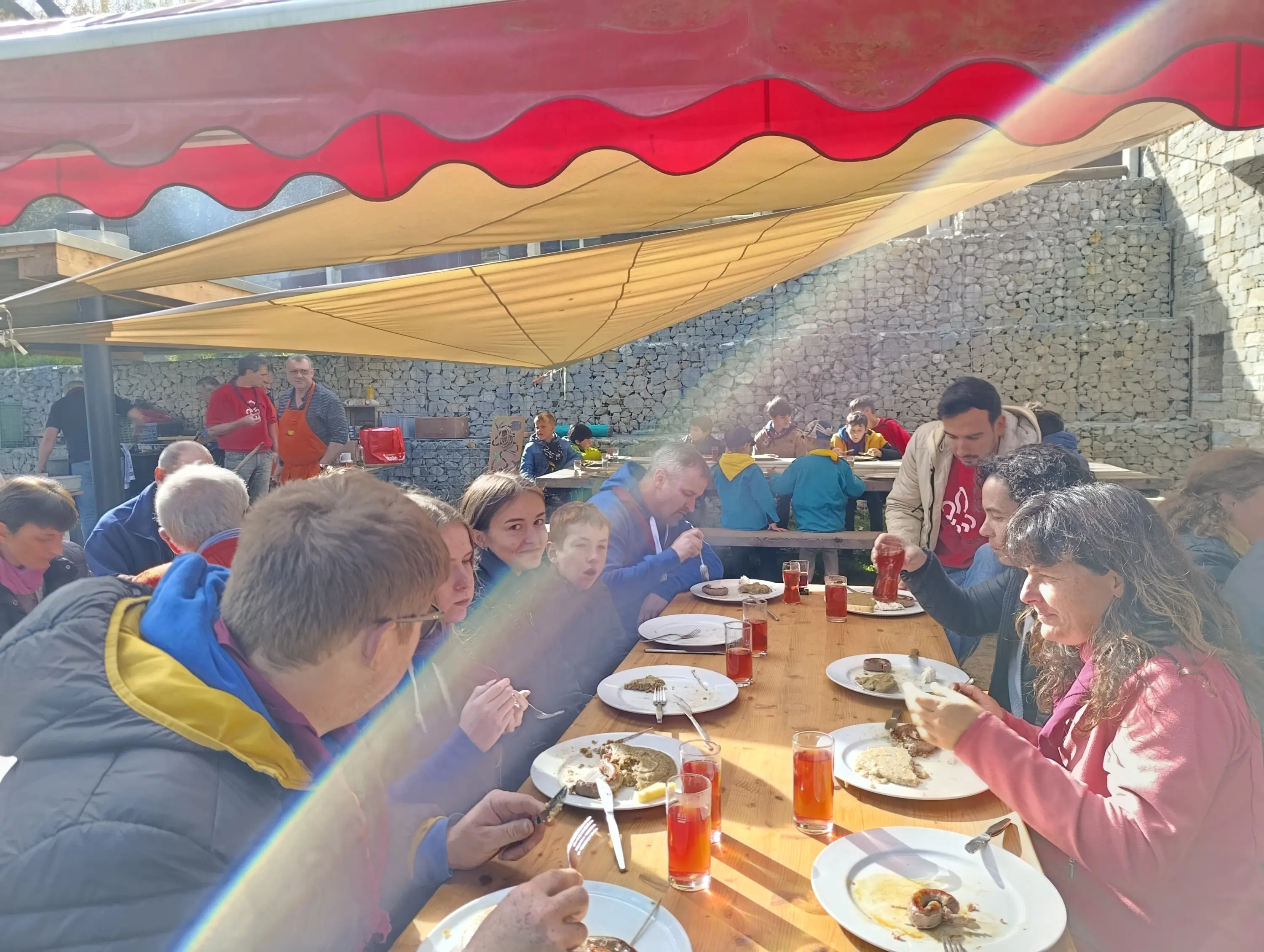 Un gruppo di persone sedute a un lungo tavolo all'aperto, mentre consumano un pasto insieme. Il sole filtra attraverso una tenda rossa, creando raggi di luce. Alcuni commensali sorridono e chiacchierano, mentre altri mangiano. Sul tavolo ci sono piatti di cibo e bevande.