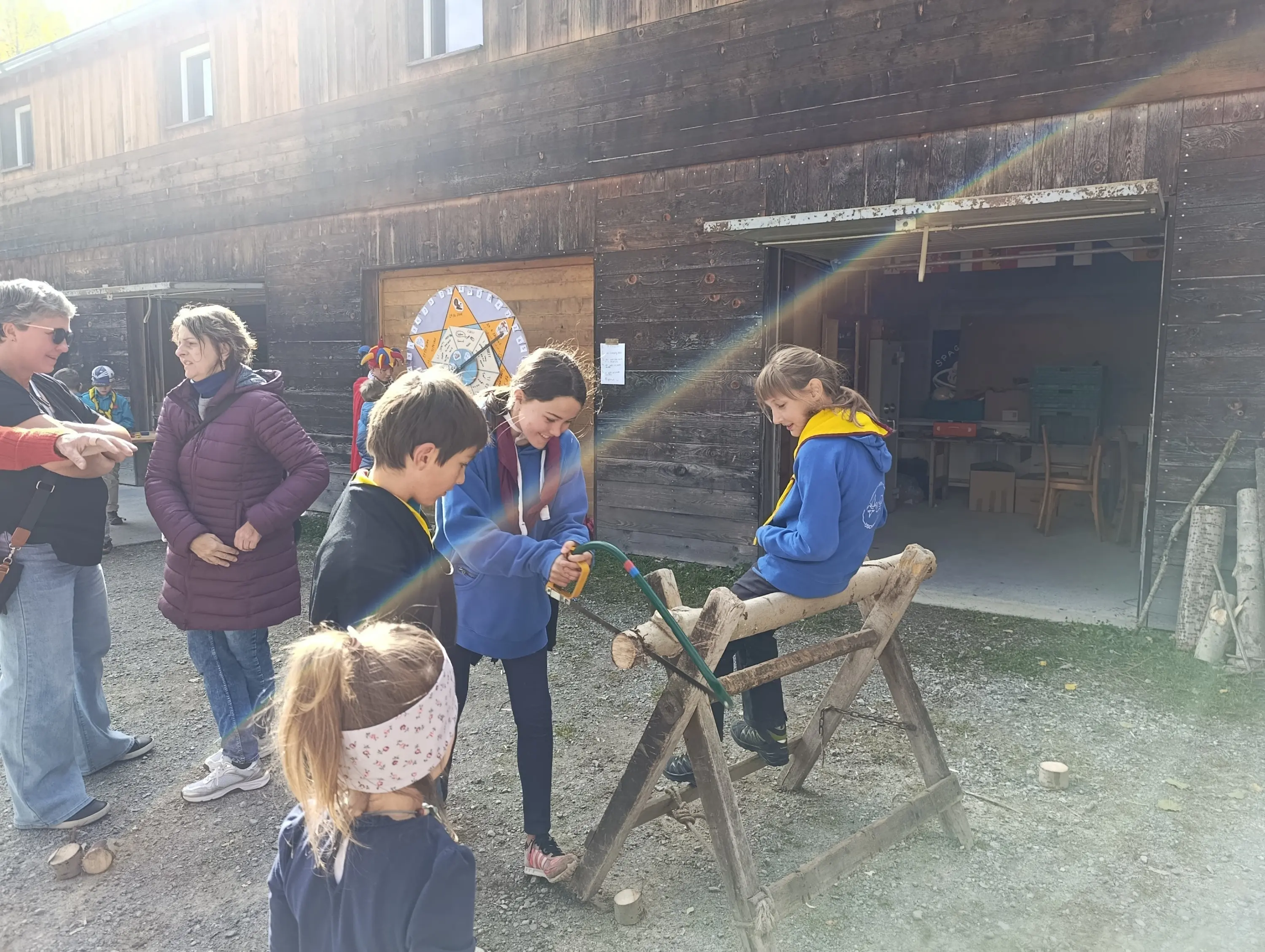 Due ragazze stanno usando un attrezzo di legno per lavorare un pezzo di legno. Altre persone osservano nei dintorni, mentre un ragazzo piccolo gioca in primo piano. Sullo sfondo si intravede un edificio in legno.