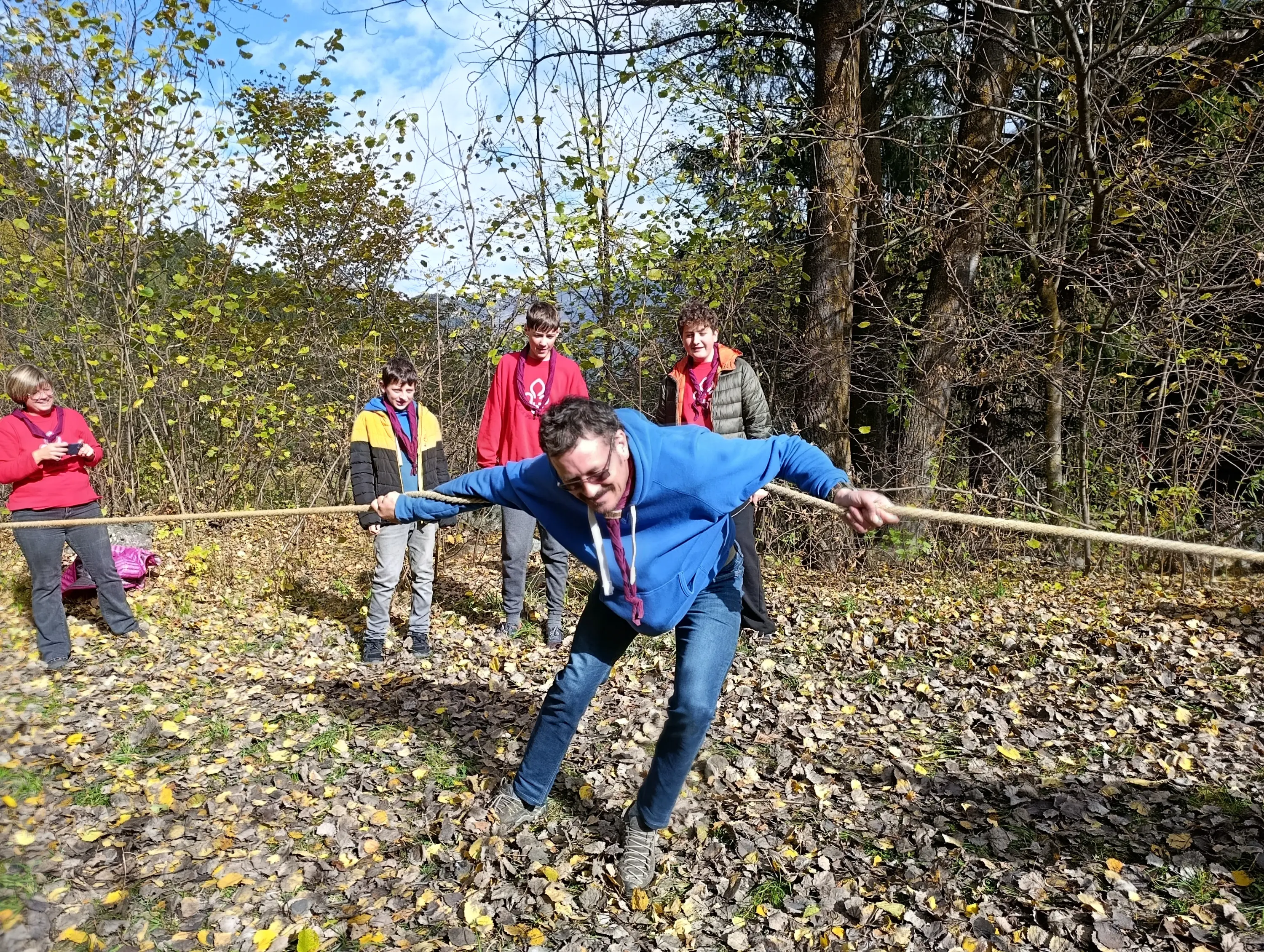 Un gruppo di persone in un bosco autunnale, con foglie gialle a terra. Un uomo in maglione blu sta tirando una corda, mentre altri ragazzi lo osservano, sorridendo e partecipando all'attività. Il cielo è parzialmente nuvoloso.