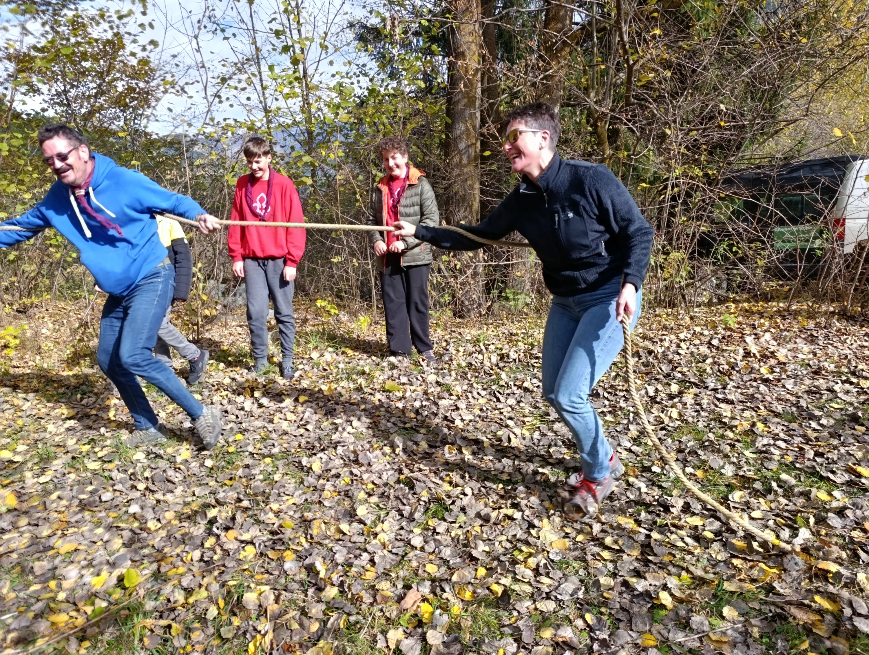 Un gruppo di persone sta giocando a tirare una corda in un prato coperto di foglie autunnali. Due persone stanno tirando in direzioni opposte, mentre altre due osservano divertite.