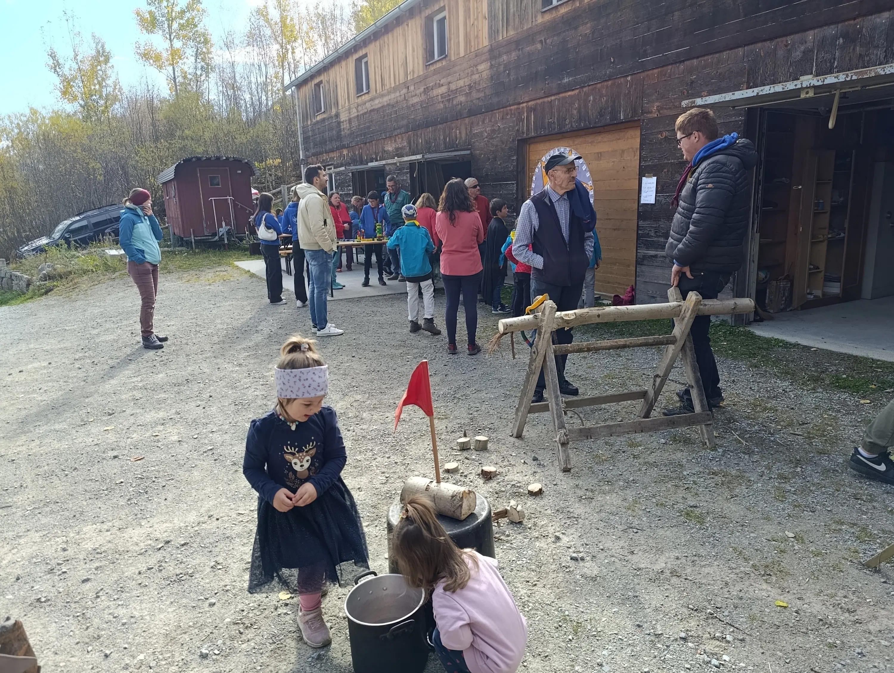 Un gruppo di persone si riunisce all'esterno di un edificio in legno, mentre alcune bambini giocano a terra. Alcuni adulti interagiscono tra loro e diversi oggetti sono visibili nel contesto, inclusi attrezzi e un tavolo. La scena è circondata da alberi con foglie che iniziano a cambiare colore.