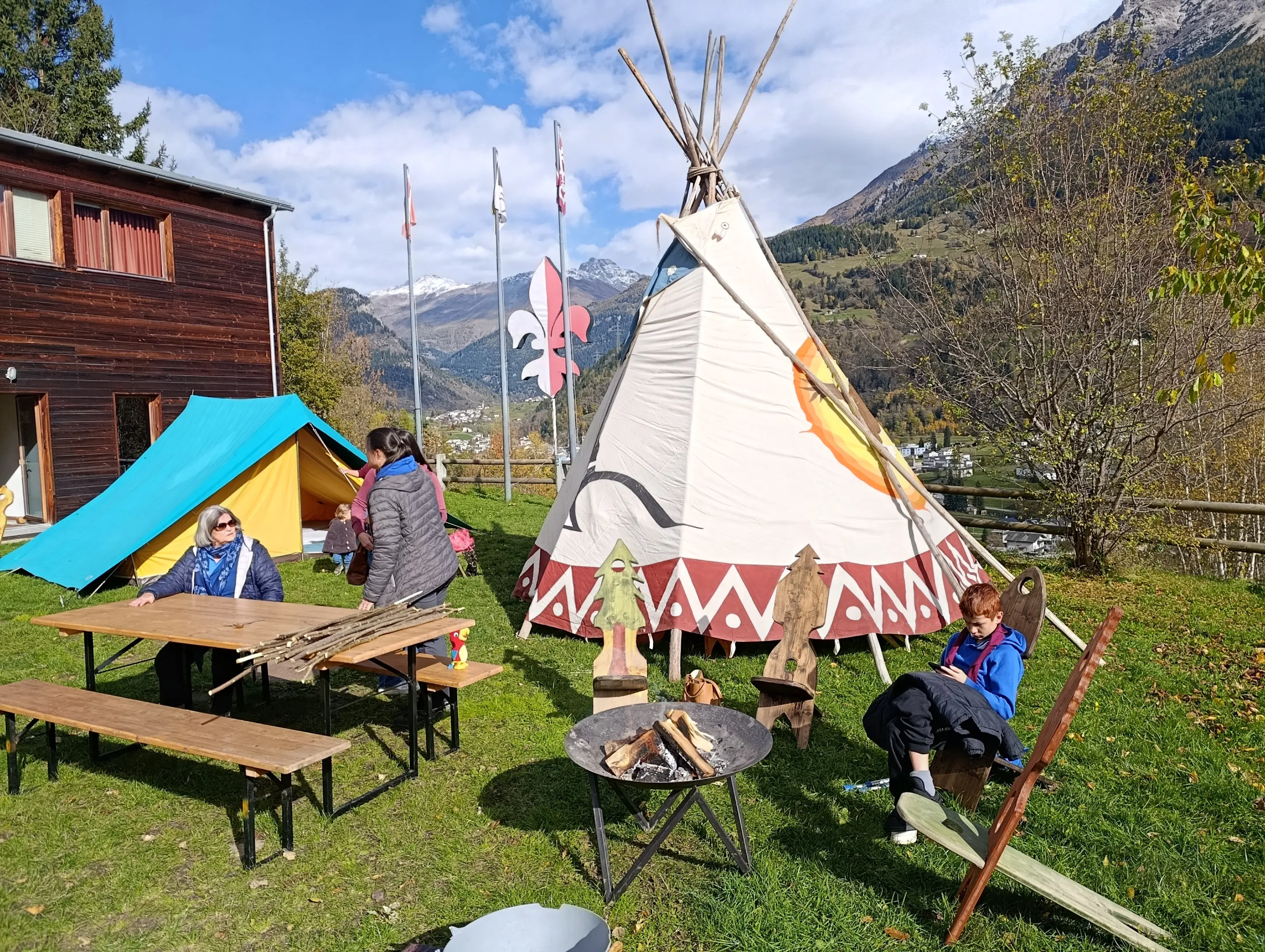 Due tende colorate e un tendone indiano sono sistemati su un prato. Un gruppo di persone si riunisce attorno a un tavolo di legno, mentre un falò acceso è al centro. Sullo sfondo, si vedono montagne e bandiere che sventolano.