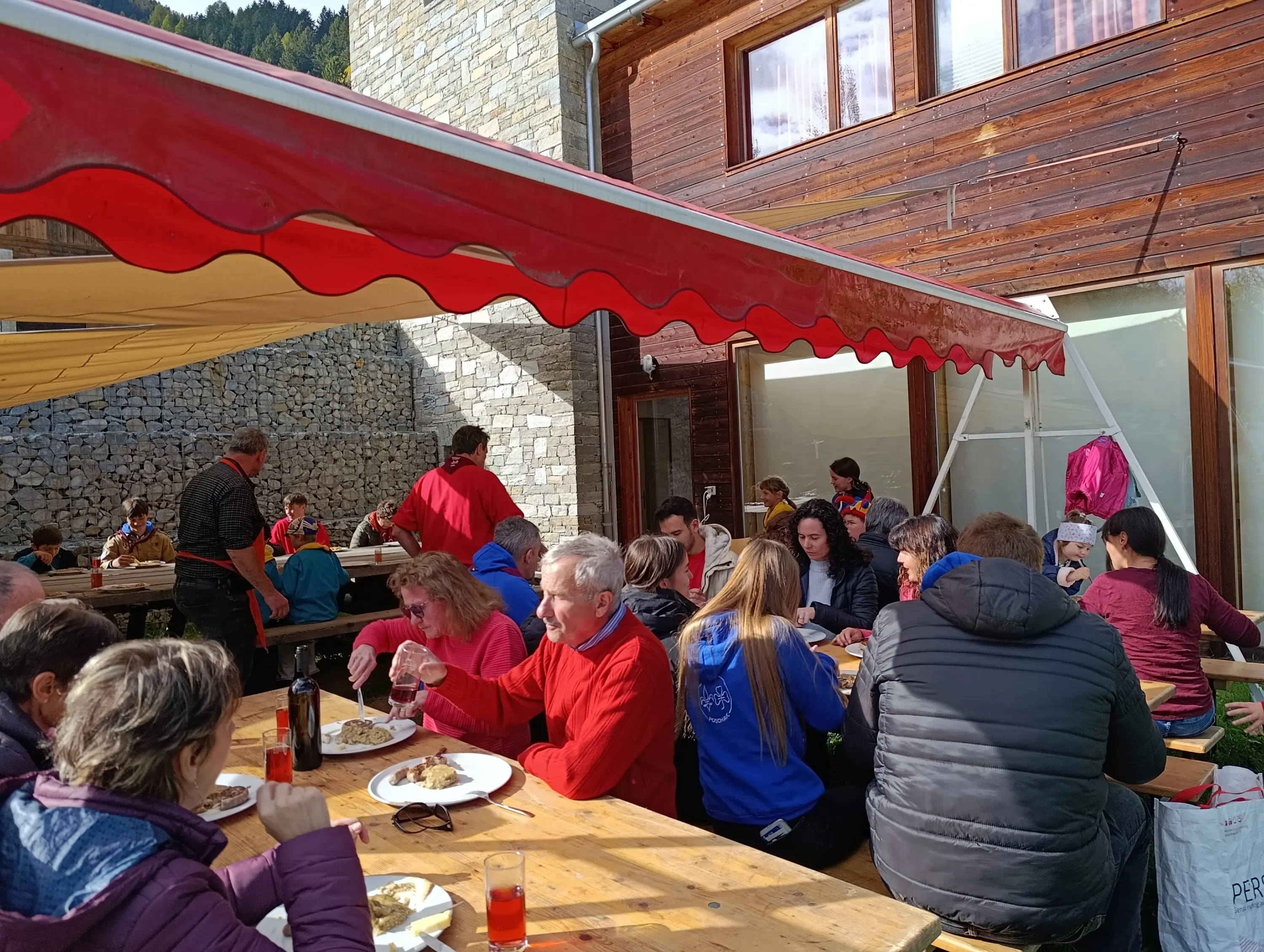 Un gruppo di persone mangia all'aperto in un rifugio di montagna, seduti a tavoli di legno sotto una tenda. Alcuni stanno conversando, mentre altri stanno mangiando piatti tipici. L'atmosfera è conviviale e rilassata, circondata da un paesaggio montano.