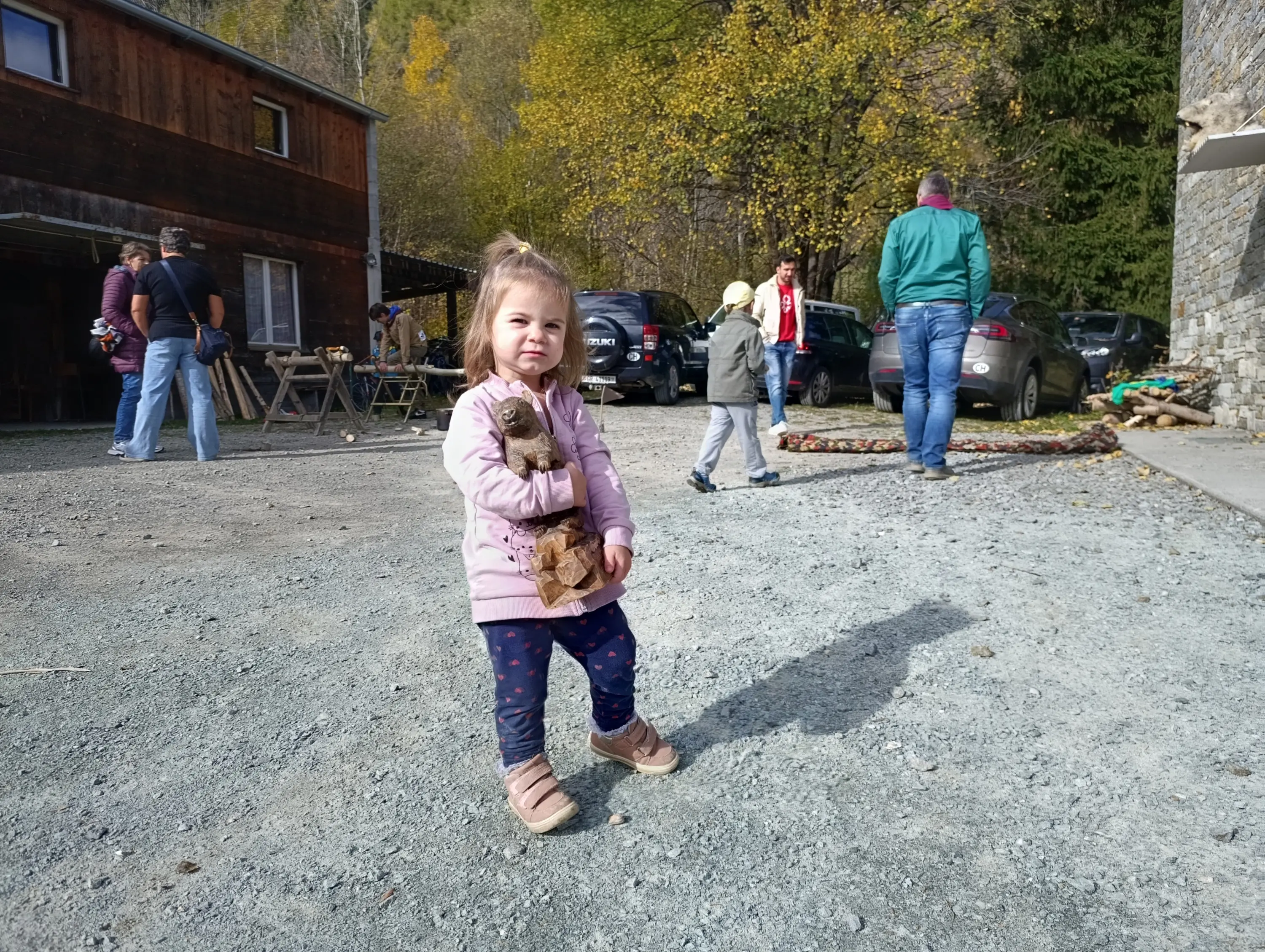 Una bambina in giacca rosa tiene in braccio un peluche. Sullo sfondo, delle persone e delle auto in un'area all'aperto circondata da alberi autunnali.