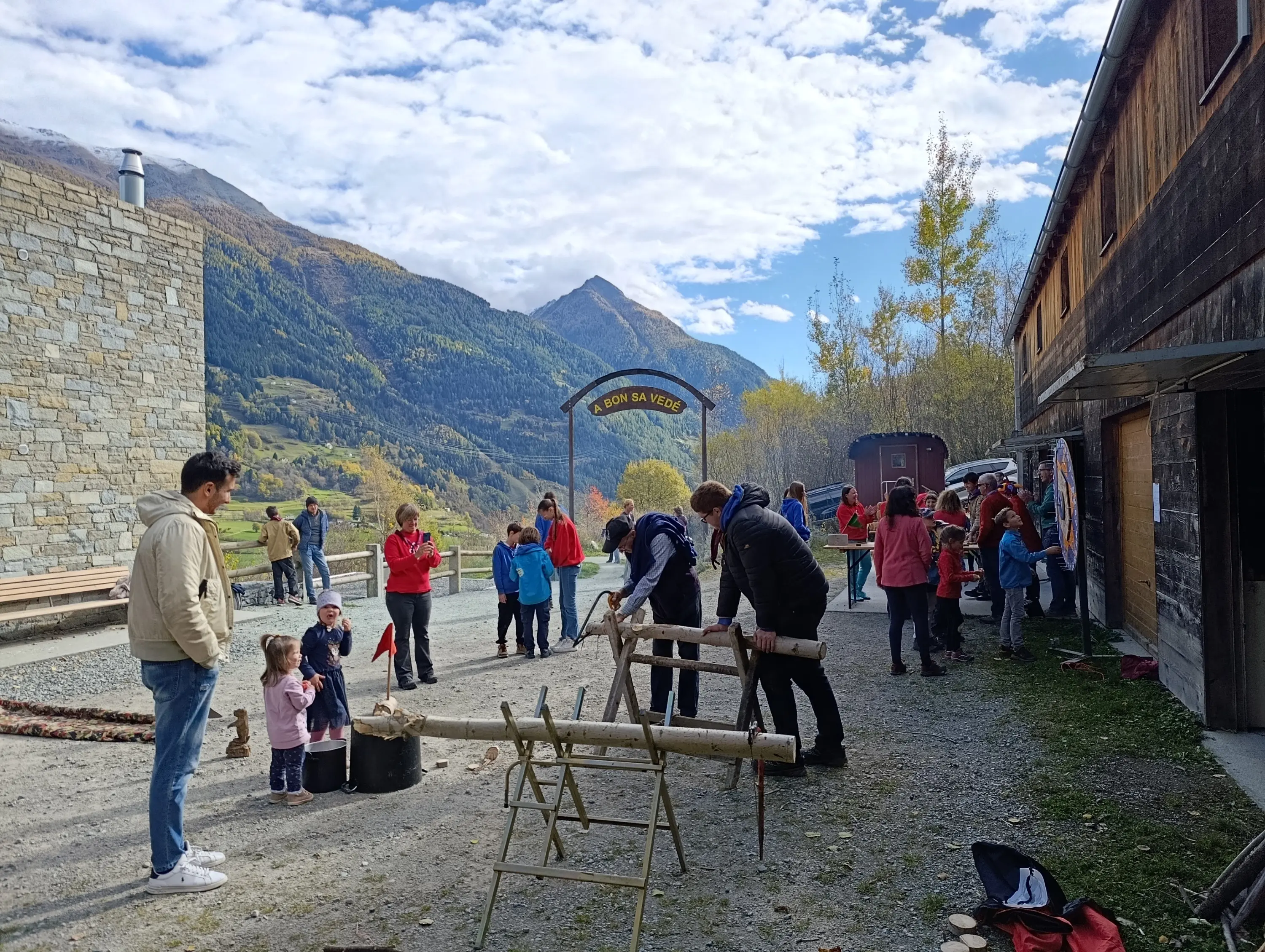 Una festa all'aperto in un ambiente montano. Bambini e adulti partecipano a varie attività, come la costruzione di strutture in legno. Alcuni membri dello staff indossano maglie rosse. Sullo sfondo, si vedono montagne e alberi autunnali. Un arco di legno segna l'ingresso di un'area chiamata "Bon