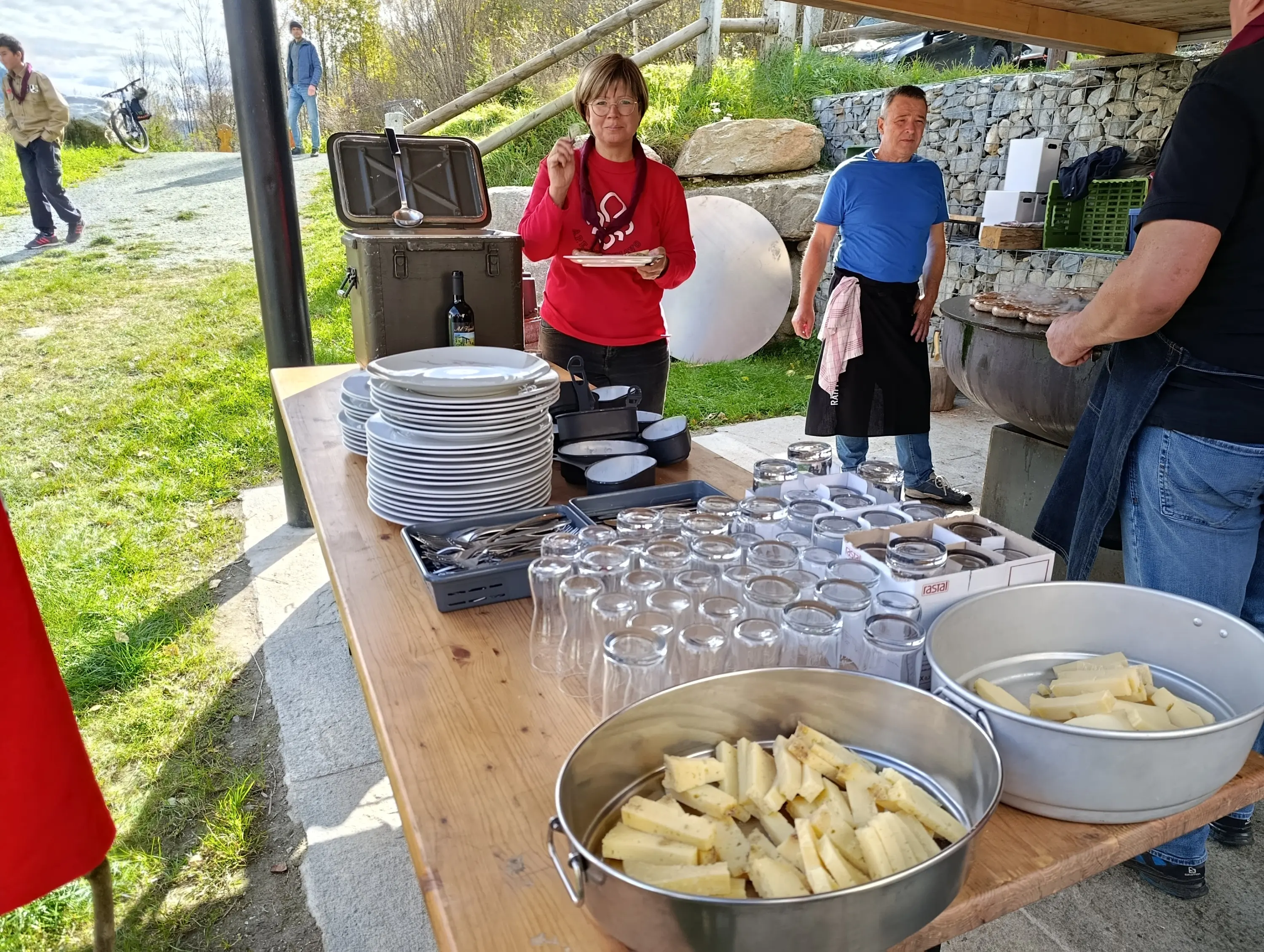 Una donna sta servendo del cibo da un tavolo all'aperto, con piatti, bicchieri e formaggi. Due uomini sono in secondo piano, mentre uno di loro guarda verso l'obiettivo. Il contesto appare informale e all'aperto.
