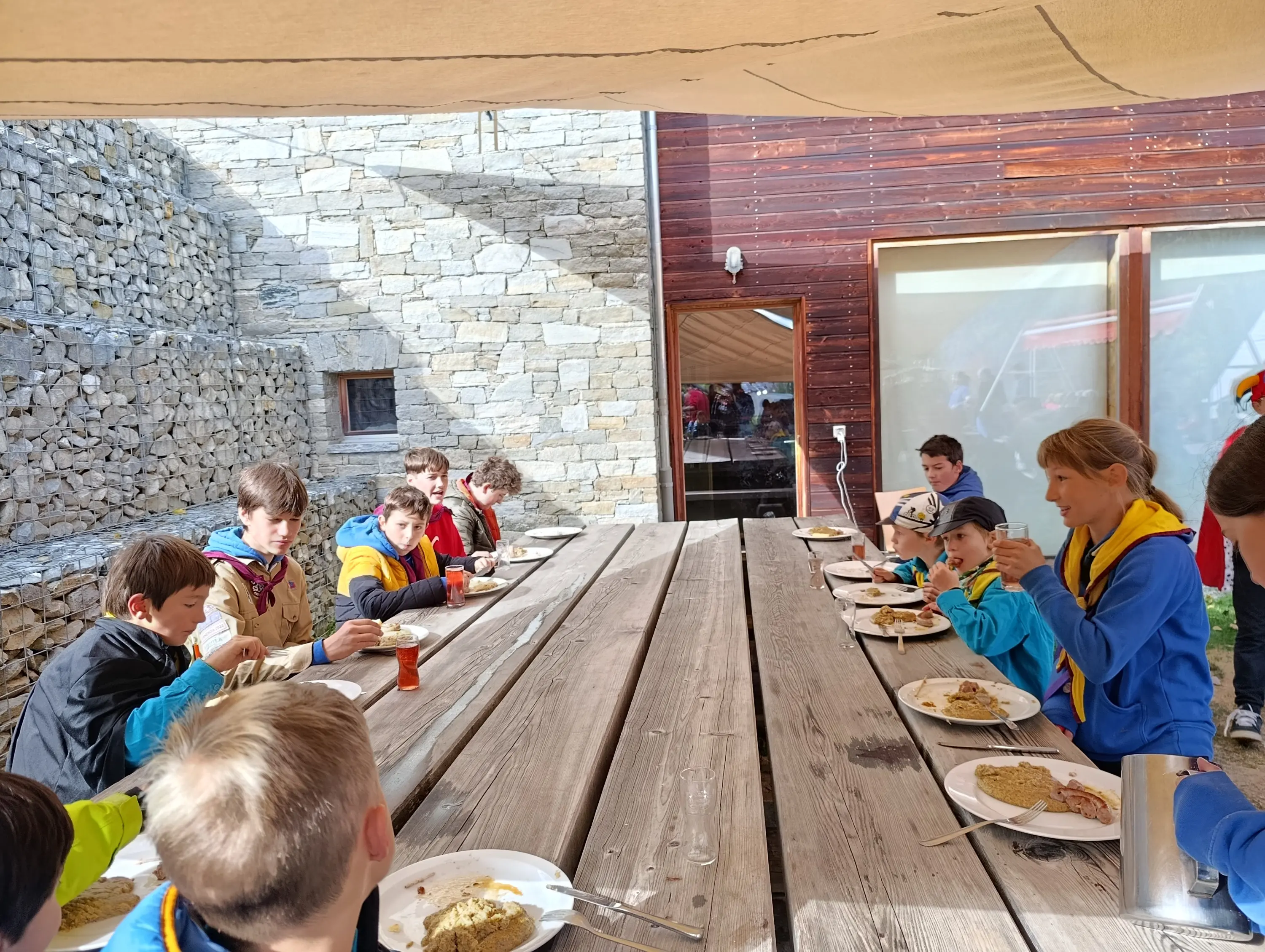 Gruppo di ragazzi seduti attorno a un lungo tavolo di legno, mentre mangiano. Alcuni di loro indossano magliette colorate. Sul tavolo ci sono piatti e posate. Lo sfondo mostra delle pareti di pietra e legno.