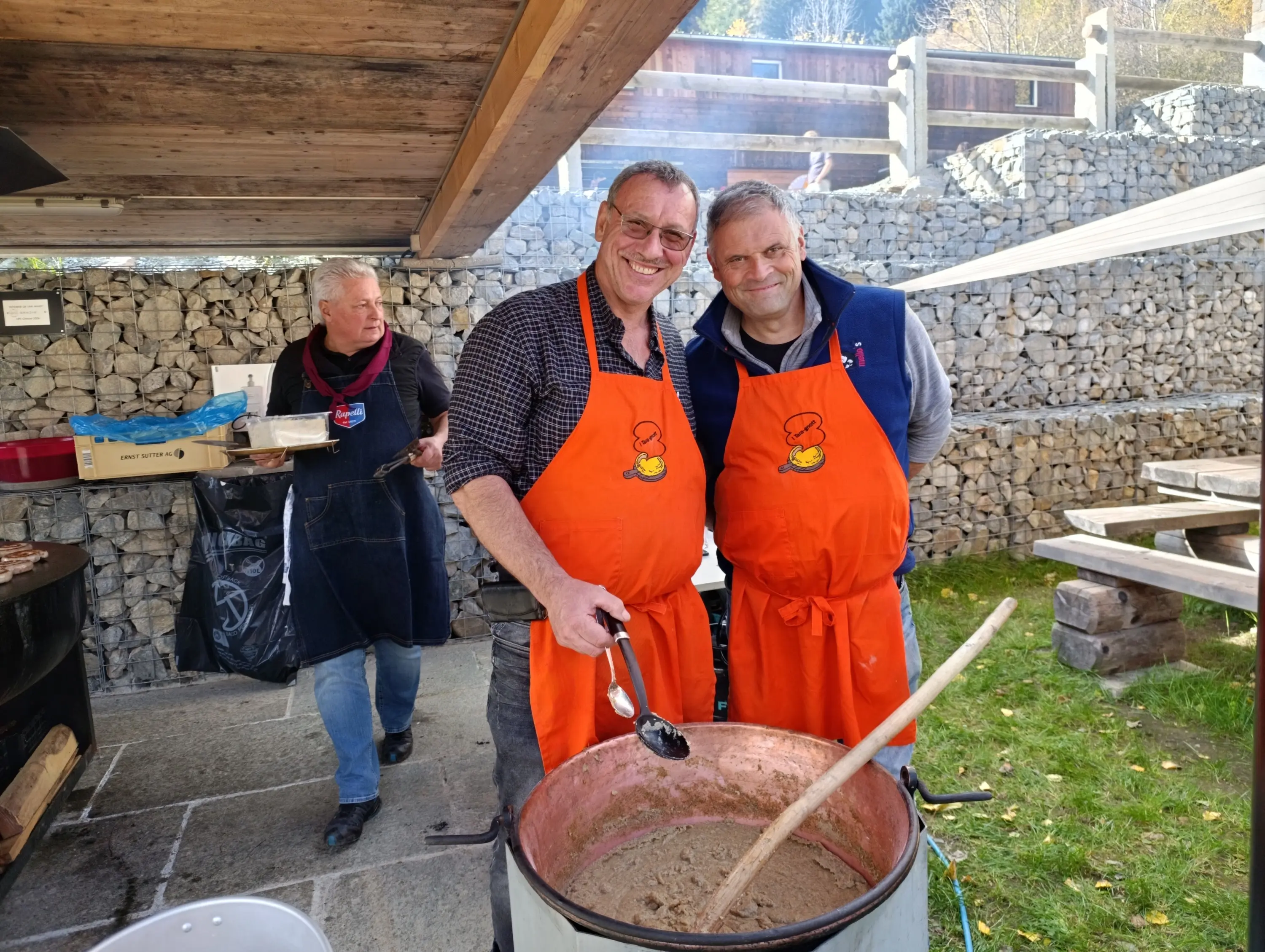 Due uomini sorridenti indossano grembiuli arancioni mentre mescolano un grande contenitore di cibo. Sullo sfondo, una donna con un grembiule nero si allontana portando un sacchetto. Un’area all'aperto con rocce e strutture in legno circosta la scena.