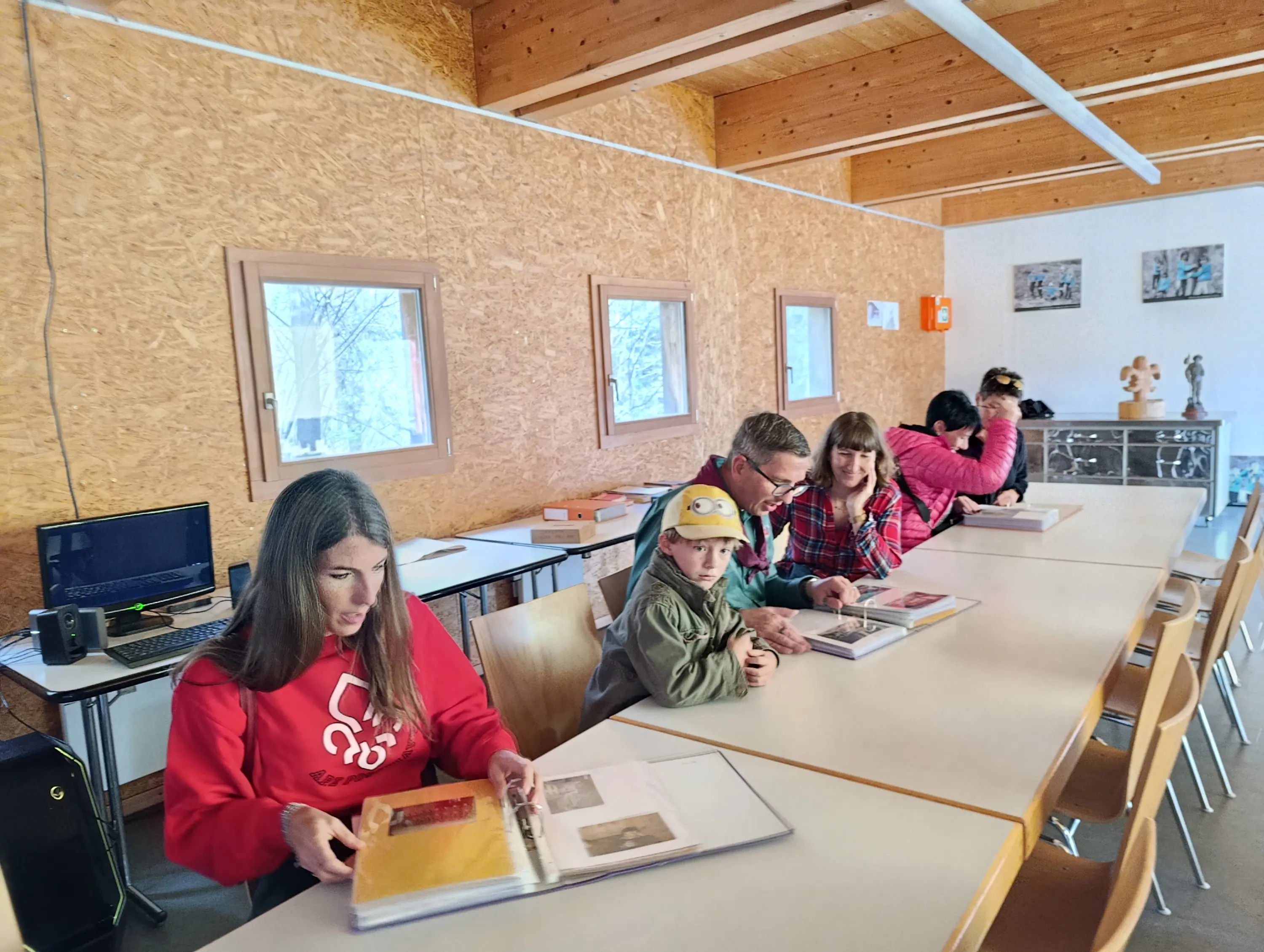 Un gruppo di persone sedute a un tavolo in una sala luminosa e accogliente, mentre sfogliano album fotografici. Due adulti e un bambino sono concentrati sulle pagine, mentre altre persone stanno interagendo più in là. L'ambiente presenta pareti di legno e finestre che lasciano entrare la luce naturale.