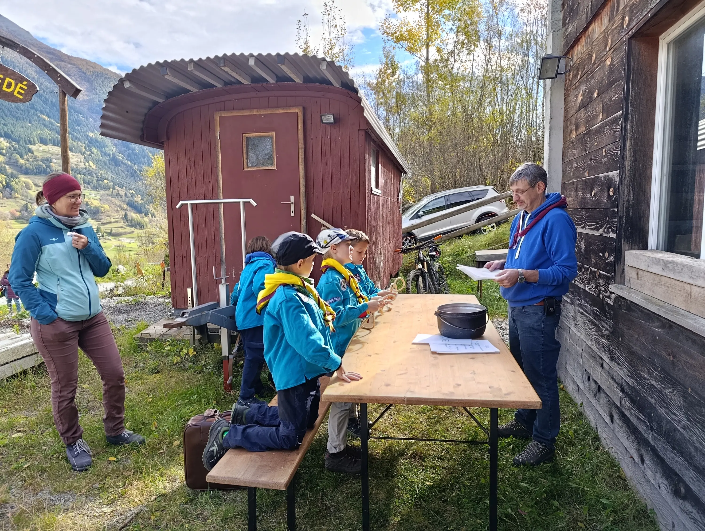 Un uomo sta parlando in piedi davanti a un tavolo all'aperto, mentre alcuni bambini ascoltano attentamente. Una donna osserva da vicino. Sullo sfondo si vedono una capanna rossa e alcuni alberi.