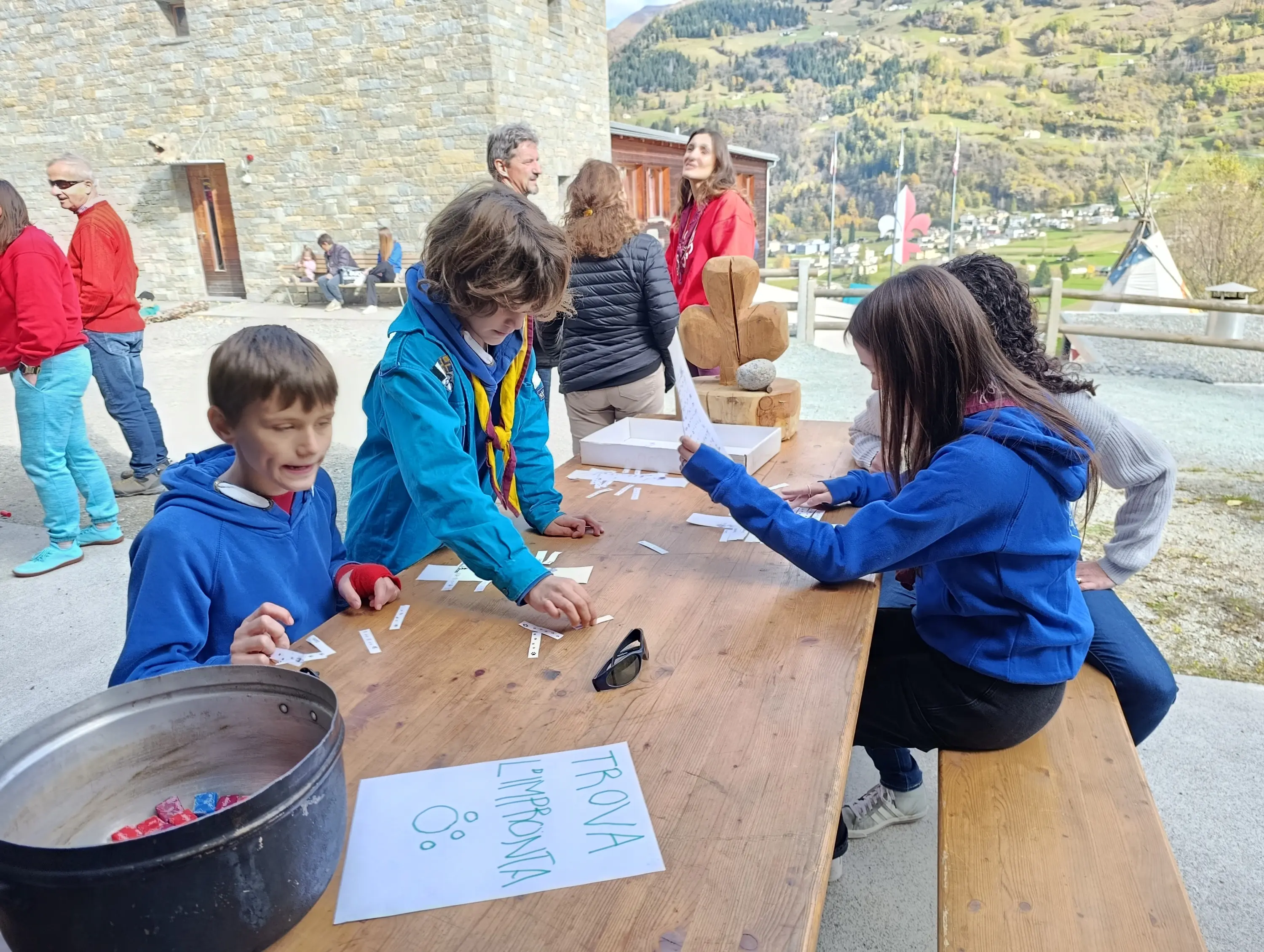 Un gruppo di bambini e adulti è riunito all'esterno di un edificio di pietra, in una zona montuosa. I bambini sono seduti a un tavolo di legno, mentre alcuni adulti osservano. Uno dei bambini scrive su un foglio con la scritta "TROVA L'IMPONDA". Si notano anche alcuni giochi e materiali sul