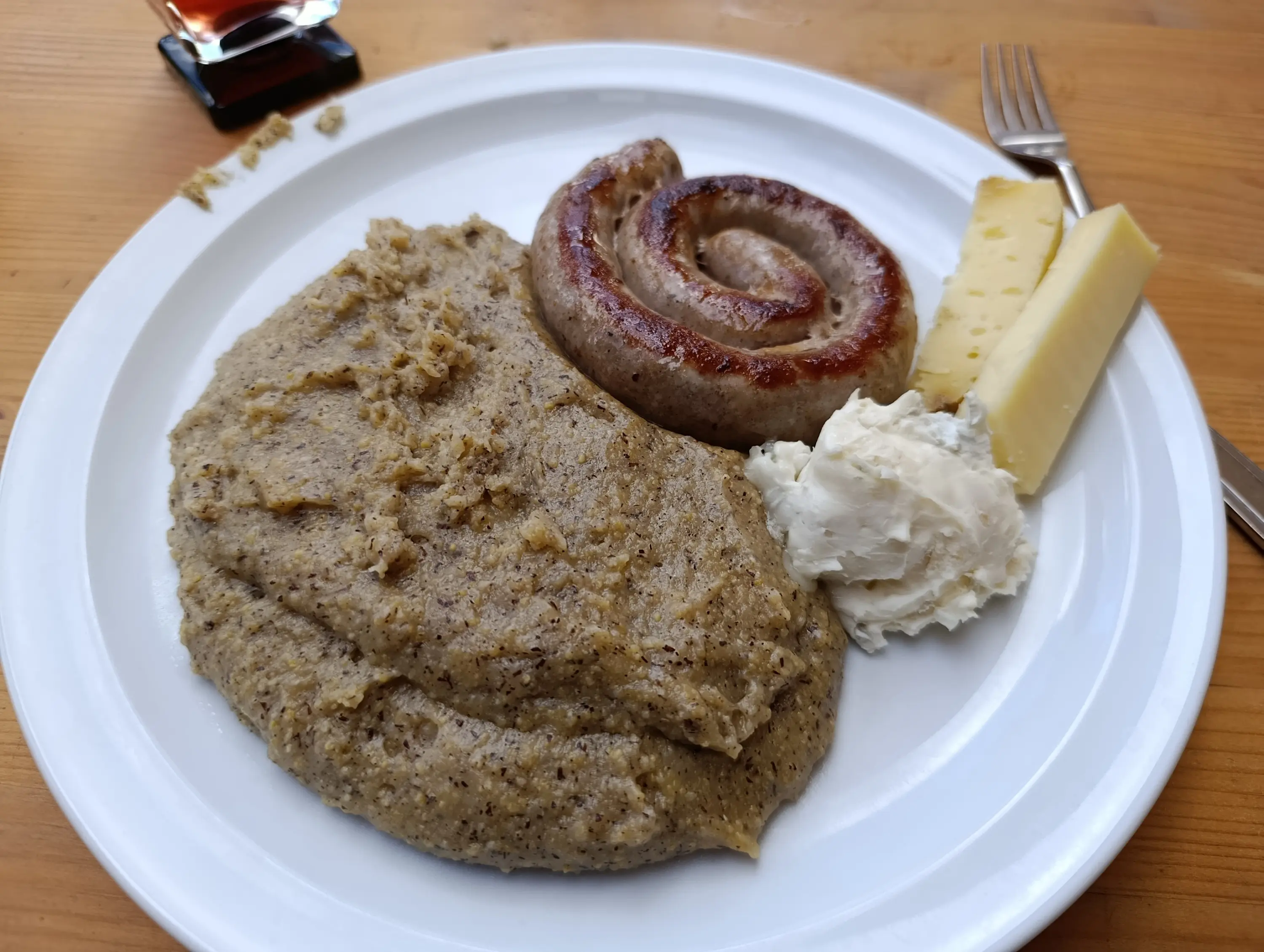 Piatto con purè di cereali scuri, salsiccia arrostita, un pezzo di formaggio e una cucchiaiata di crema. Fondo di legno chiaro.