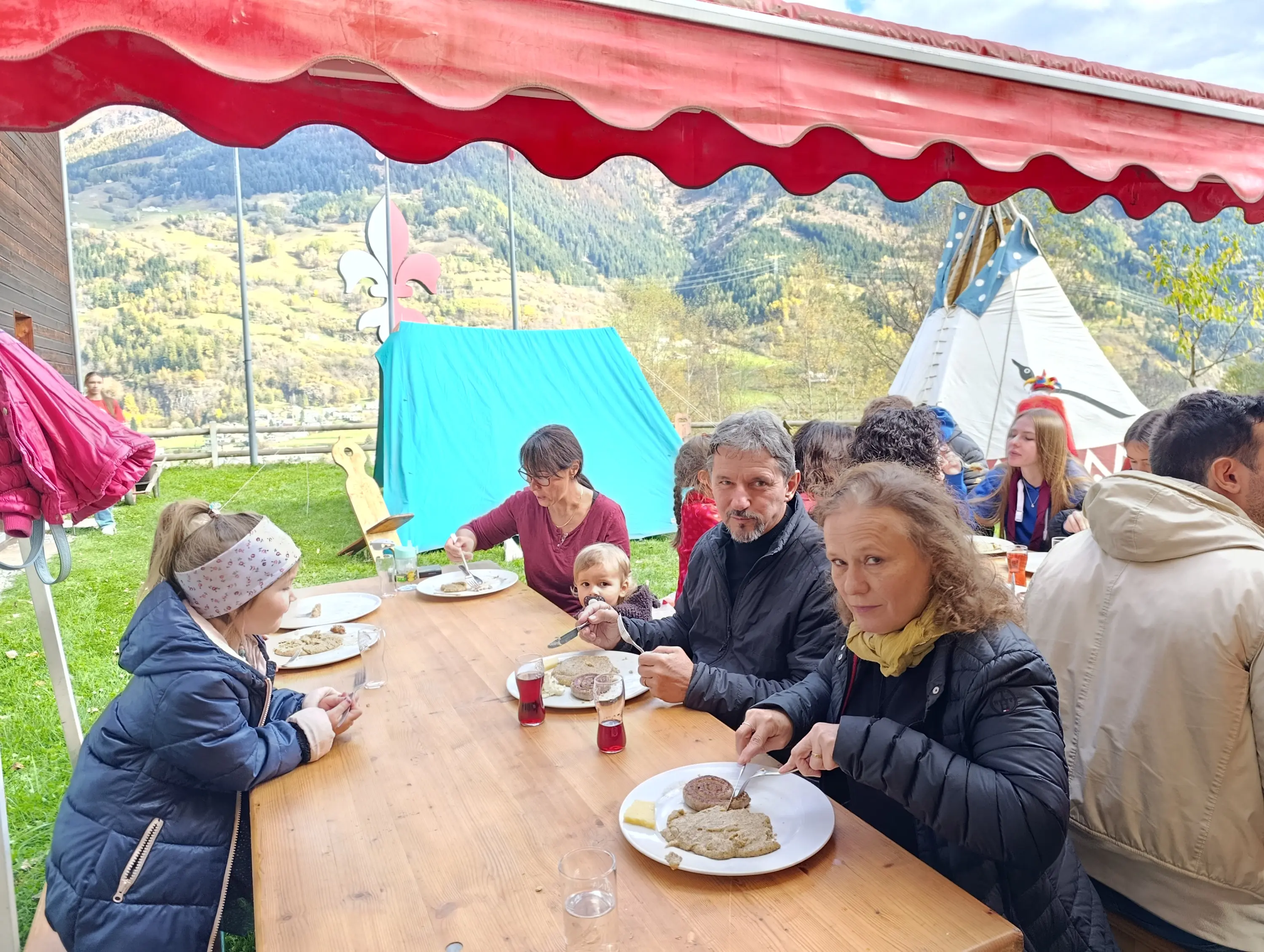 Persone sedute a un tavolo all'aperto, mentre mangiano piatti di cibo. Si vedono adulti e bambini, in un ambiente montano con tende sullo sfondo. È una giornata soleggiata e ci sono decorazioni colorate.