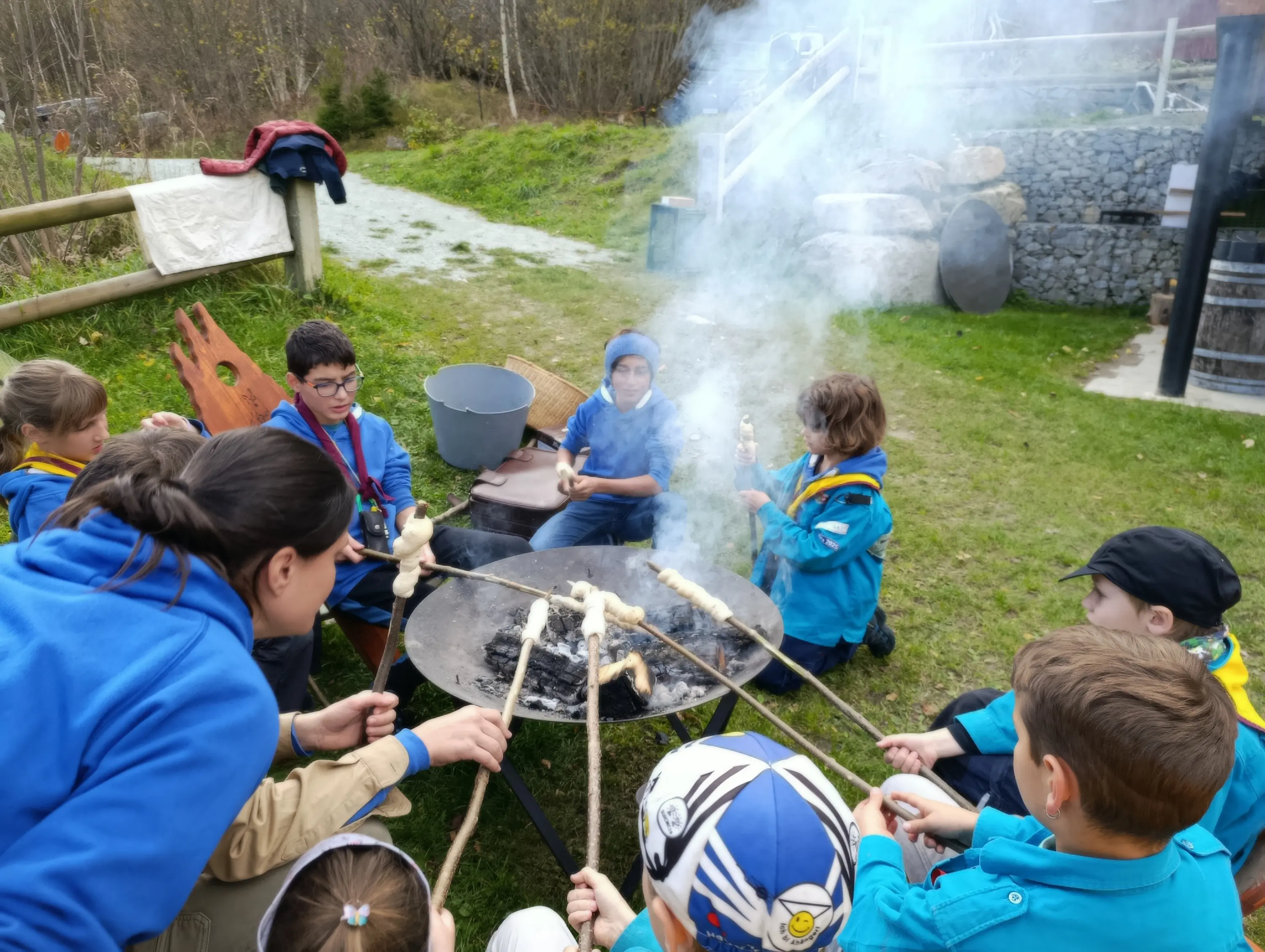 Un gruppo di bambini e adolescenti in uniformi blu si riunisce attorno a un falò, mentre cucinano pane su bastoni. Il fumo si alza nell'aria e l'ambiente circostante è naturale e verde.