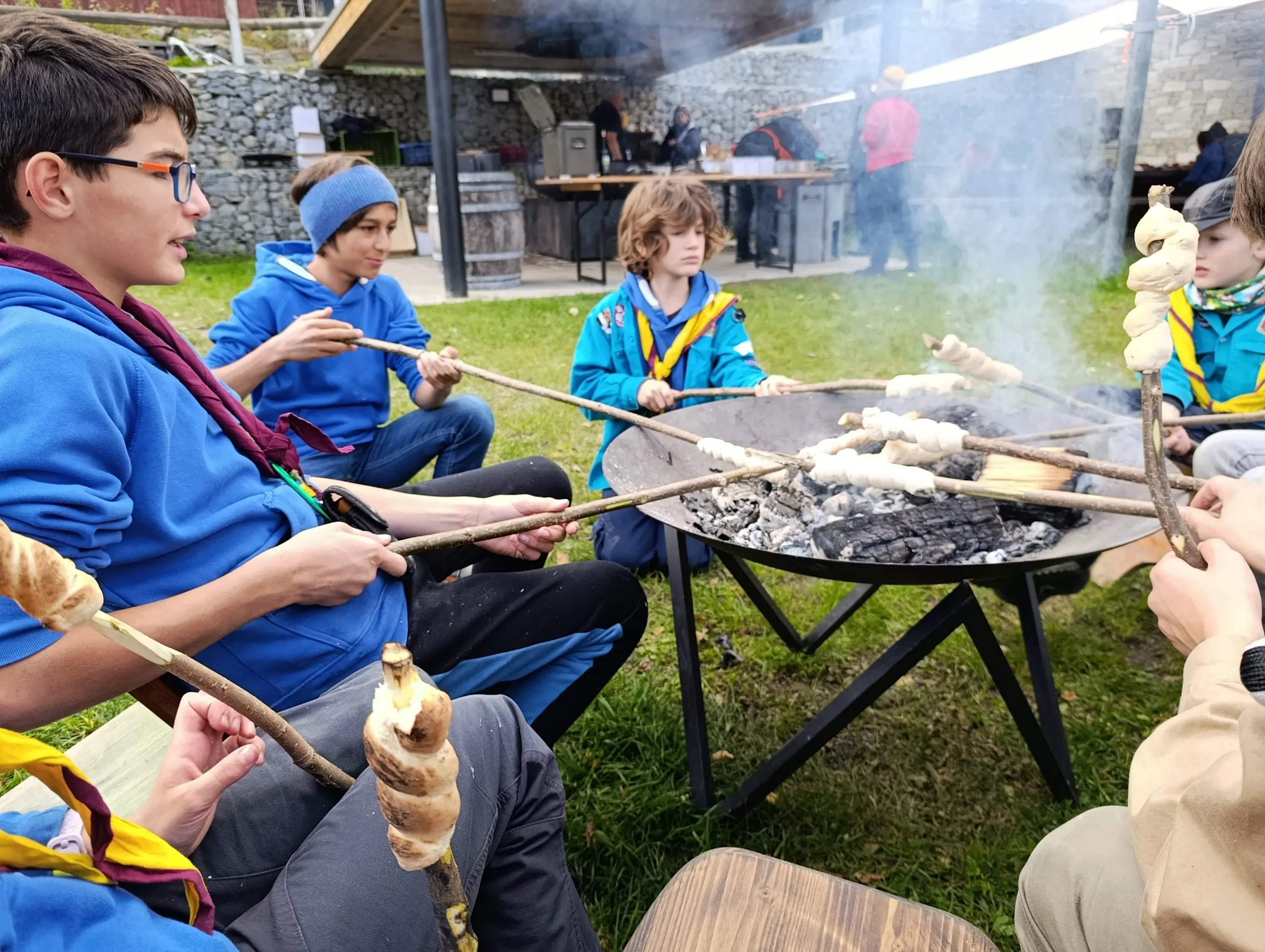Giovani scout siedono attorno a un fuoco, mentre arrostiscono impasti su lunghi bastoni. Alcuni indossano maglioni blu e sciarpe colorate. Sullo sfondo si vede una struttura con altre persone e un'atmosfera conviviale.
