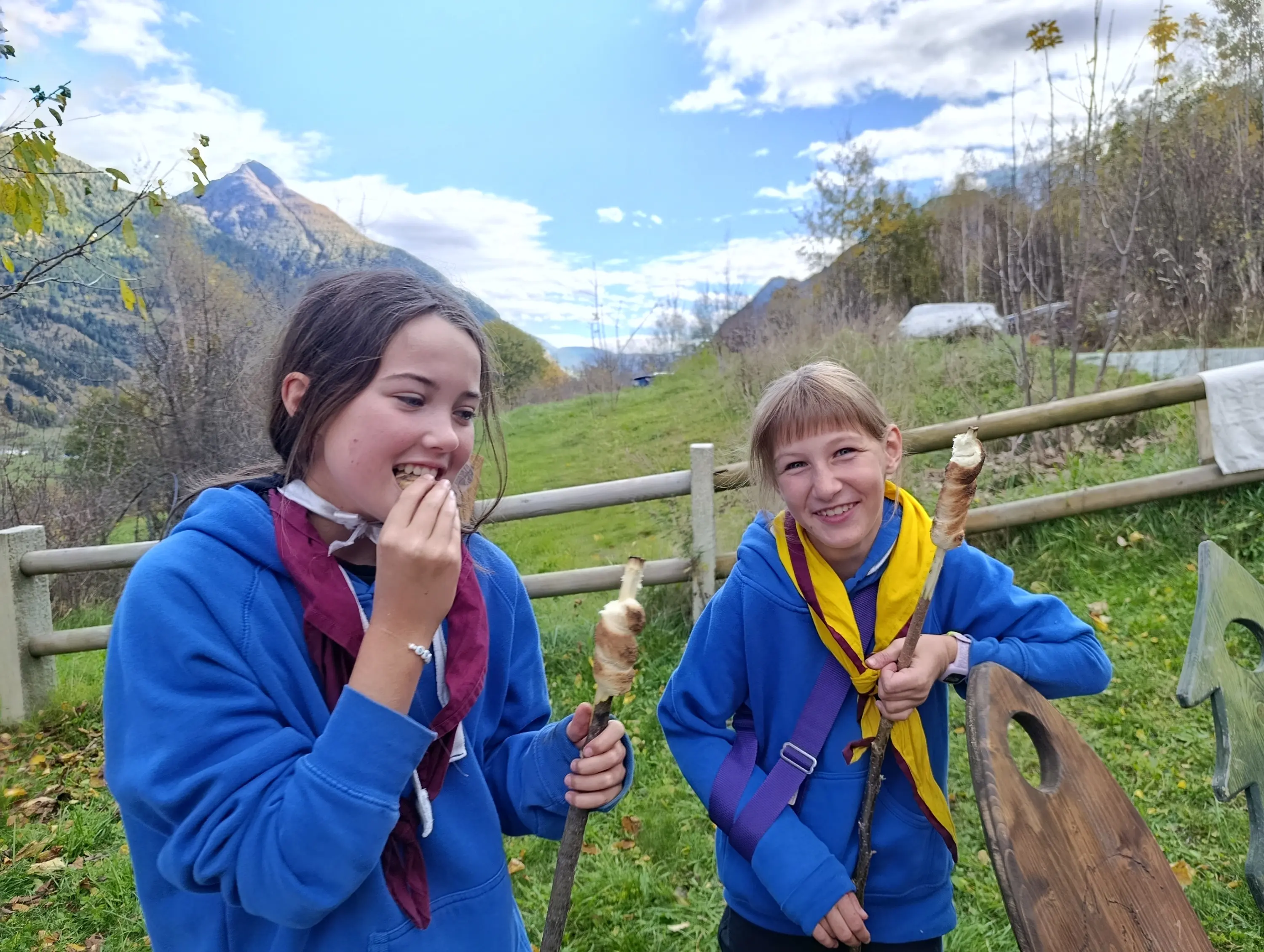 Due ragazze sorridenti in maglioni blu si godono un dolce mentre si trovano all'aperto, circondate da un paesaggio montano. Sullo sfondo, ci sono alberi e colline.