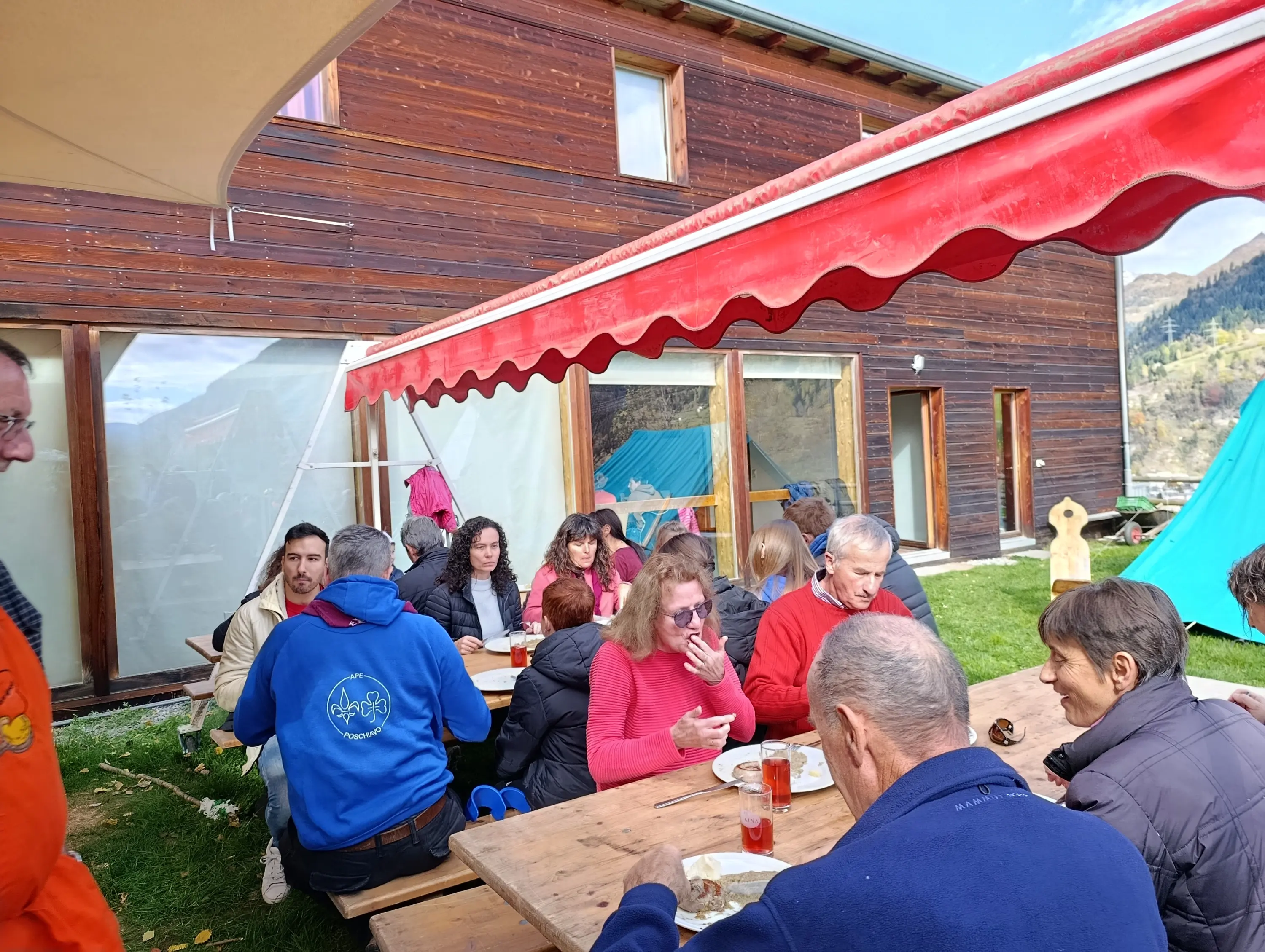Un gruppo di persone sedute a un tavolo all'aperto, mentre consumano un pasto insieme. Sullo sfondo si vede una grande casa di legno e alcune tende montate nel giardino.